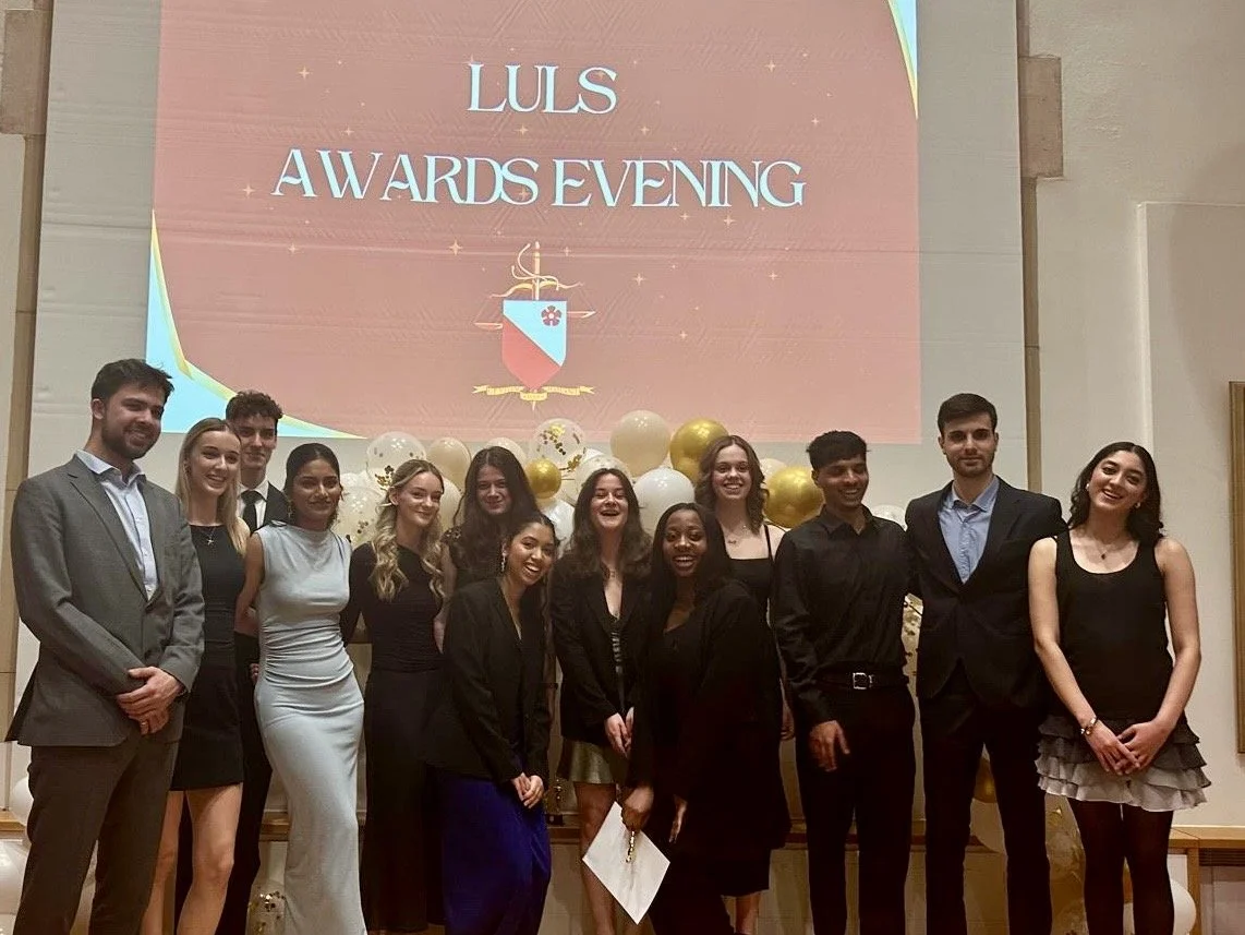 Group of young adults at LULS awards evening in formal attire, standing in front of a decorated stage with balloons and a large screen displaying the event name.