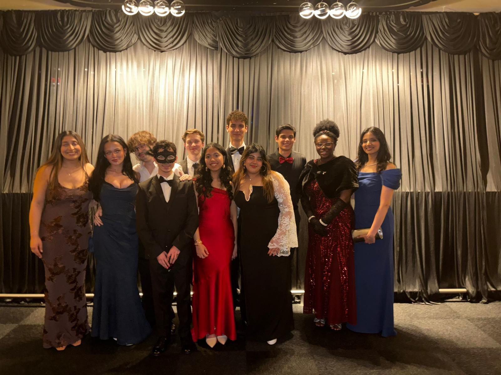 Group of people dressed in formal attire standing on stage with a curtain backdrop, likely at a prom or formal event.
