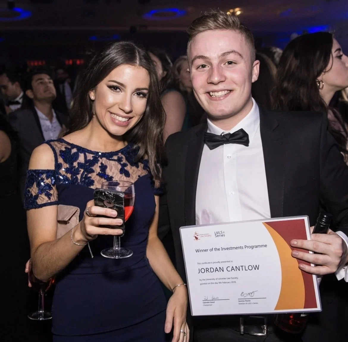 A young woman in a navy dress holding a glass of red wine standing next to a smiling young man in a tuxedo holding a certificate at an award event.