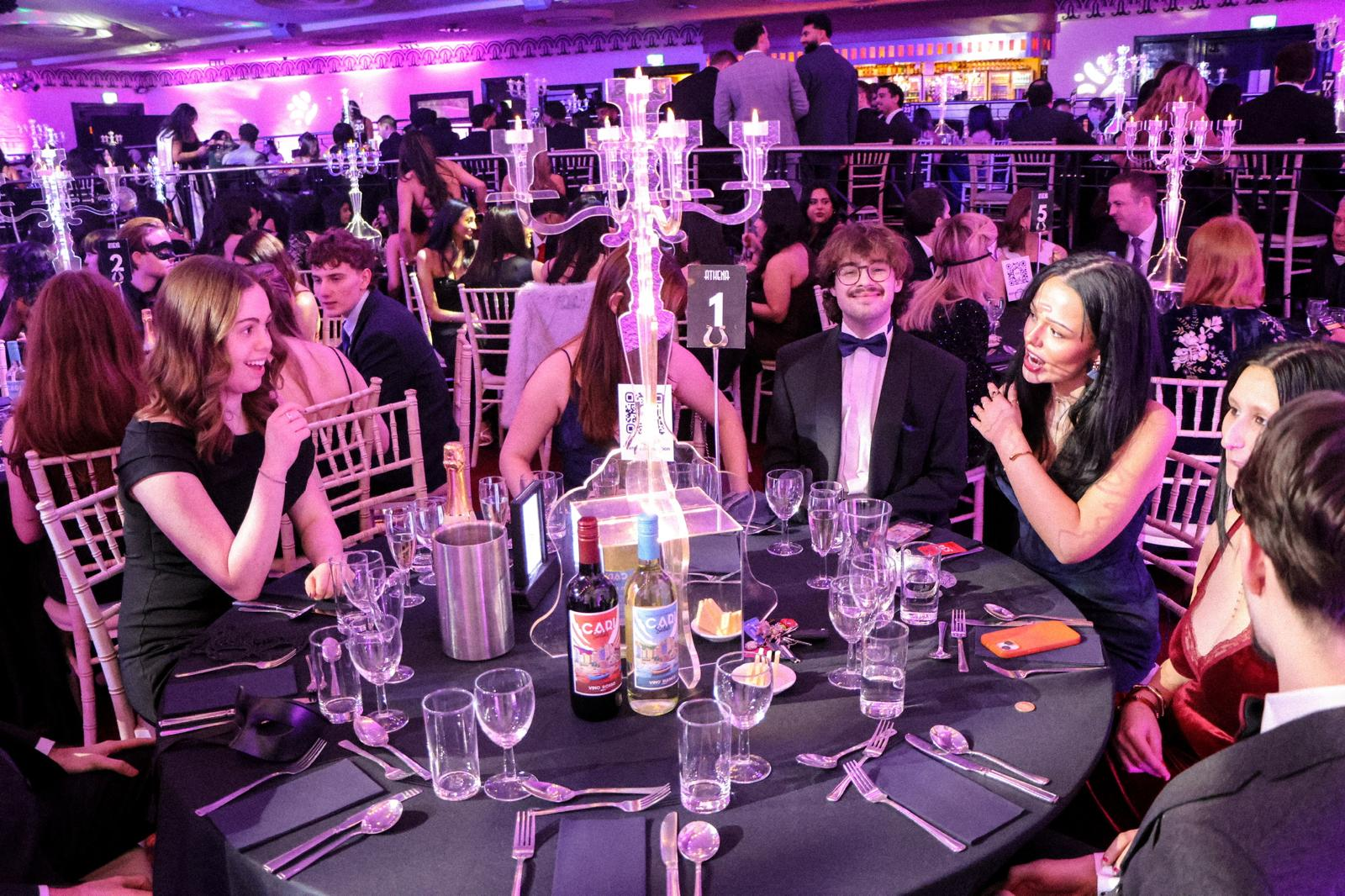 People sitting at a formal dinner table with black tablecloths, surrounded by glasses, bottles of alcohol, and a candelabra centerpiece, at an event or party with purple lighting and many guests in formal attire.