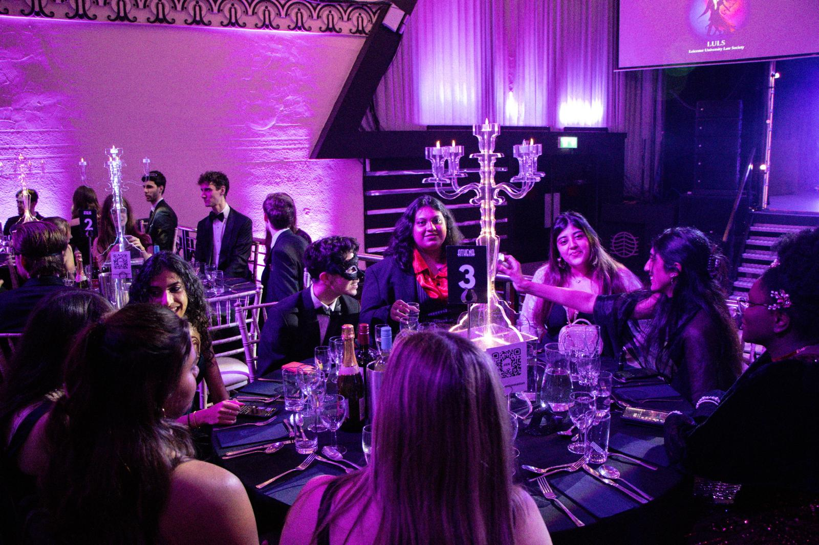A formal dinner event with guests seated at a round table decorated with a large candelabra centerpiece and bottles of wine and champagne. The scene is illuminated with purple lighting, and some guests are wearing masks. There is a large screen in th