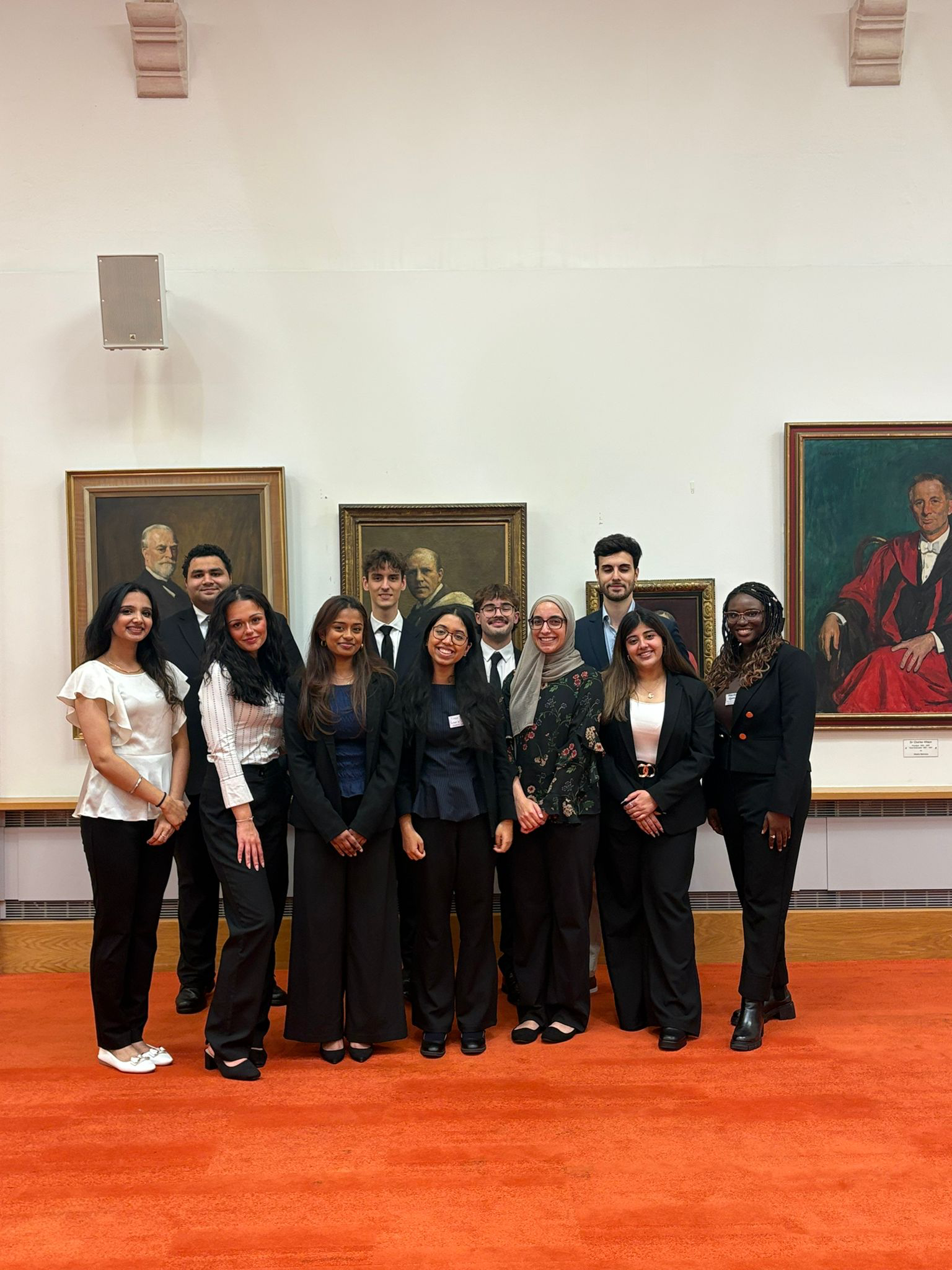 Group of young adults in formal attire posing for a photo at an art gallery with classical style portraits on the wall.