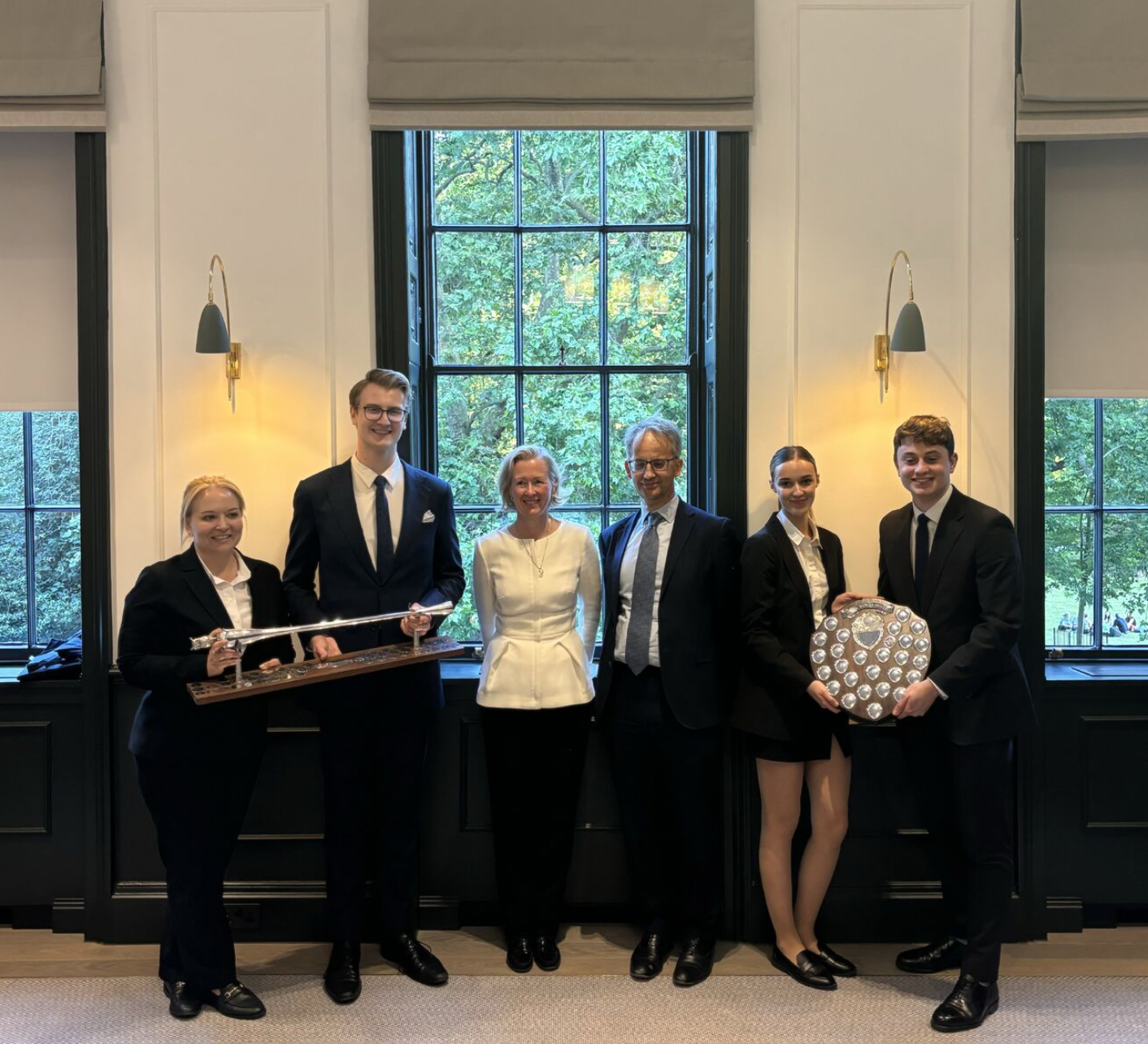 Group of six people standing indoors in front of large windows with greenery outside. Two young people are holding shields, one with a sword and another with a trophy or shield with multiple small shields. The group includes three women and three men