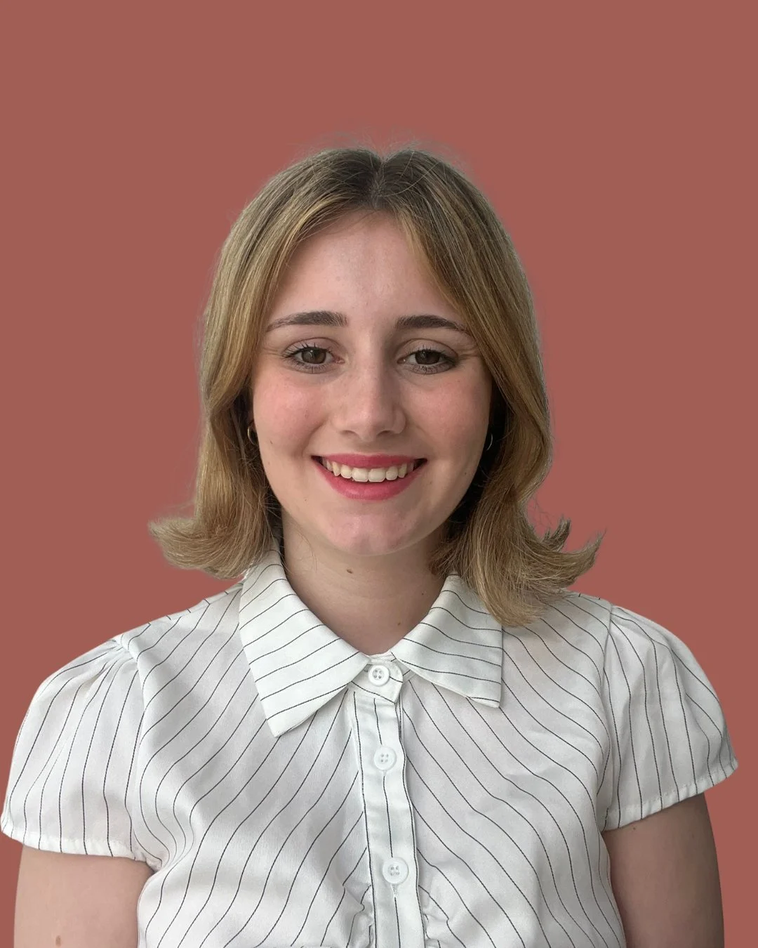 A young woman with shoulder-length light brown hair smiling, wearing a white shirt with thin black stripes, against a plain muted pink background.