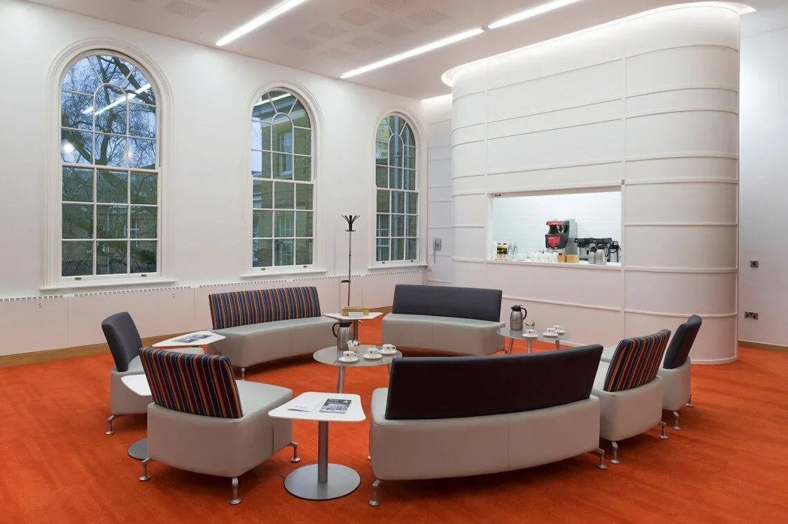 Modern lounge area with curved gray sofas, striped upholstered chairs, small round tables with coffee cups and papers, large arched windows, white walls, and an orange carpet. A coffee machine is visible through a pass-through window in a curved whit