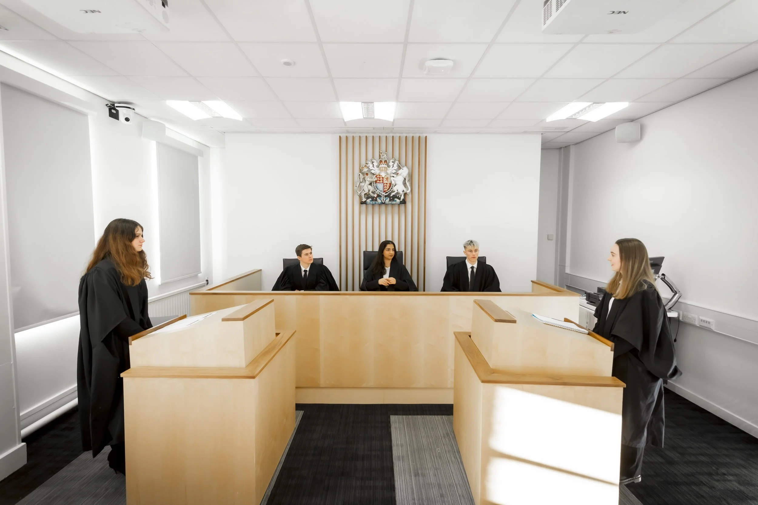 A courtroom with five young people dressed in black robes, sitting behind a wooden bench. Two women stand on opposite sides of the room, facing each other. The room has white walls, ceiling lights, and a coat of arms or official emblem on the wall be