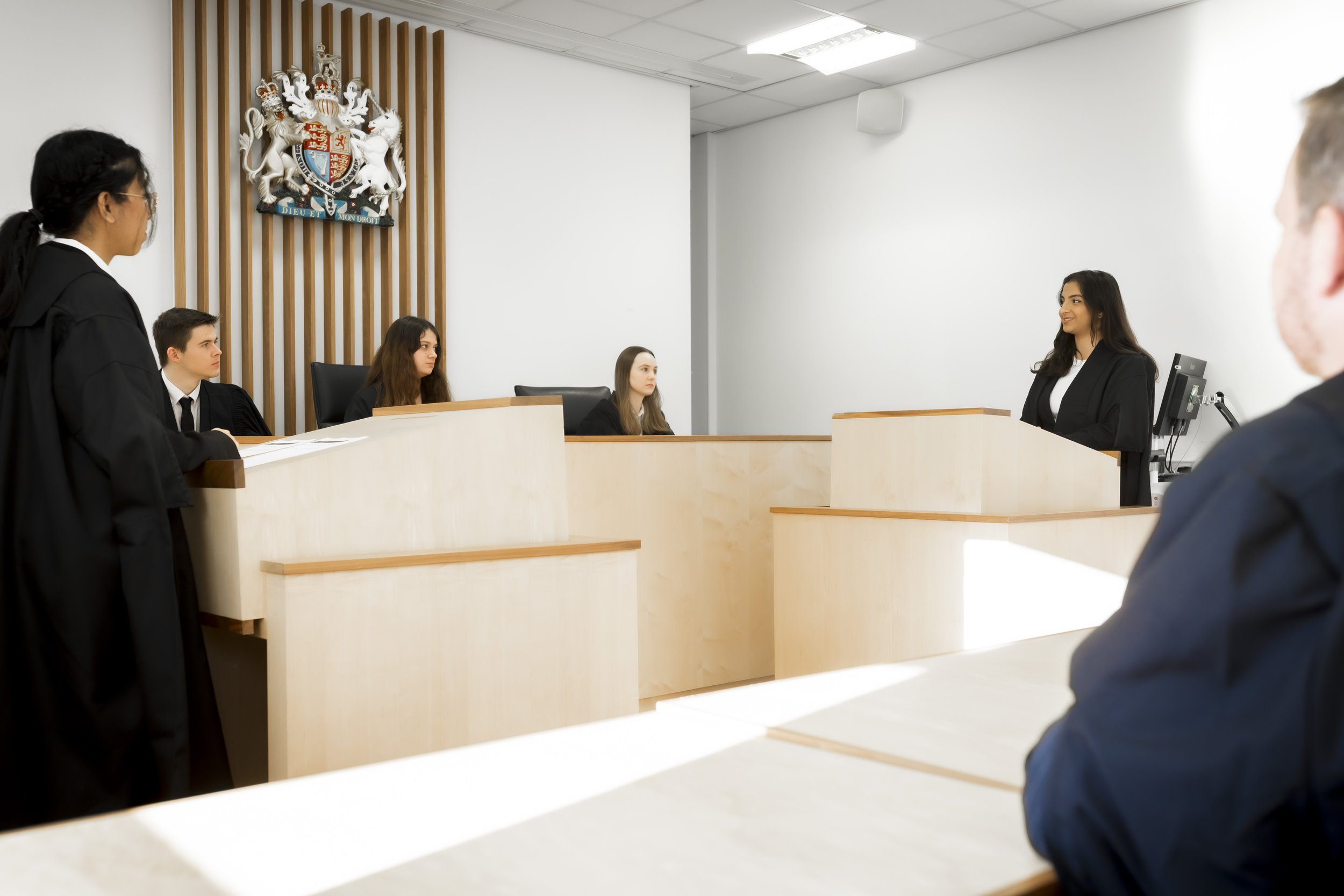 A woman in a black suit is standing at a wooden podium, speaking in a courtroom with four seated individuals, including a judge and three young adults. The courtroom features a coat of arms on the wall.