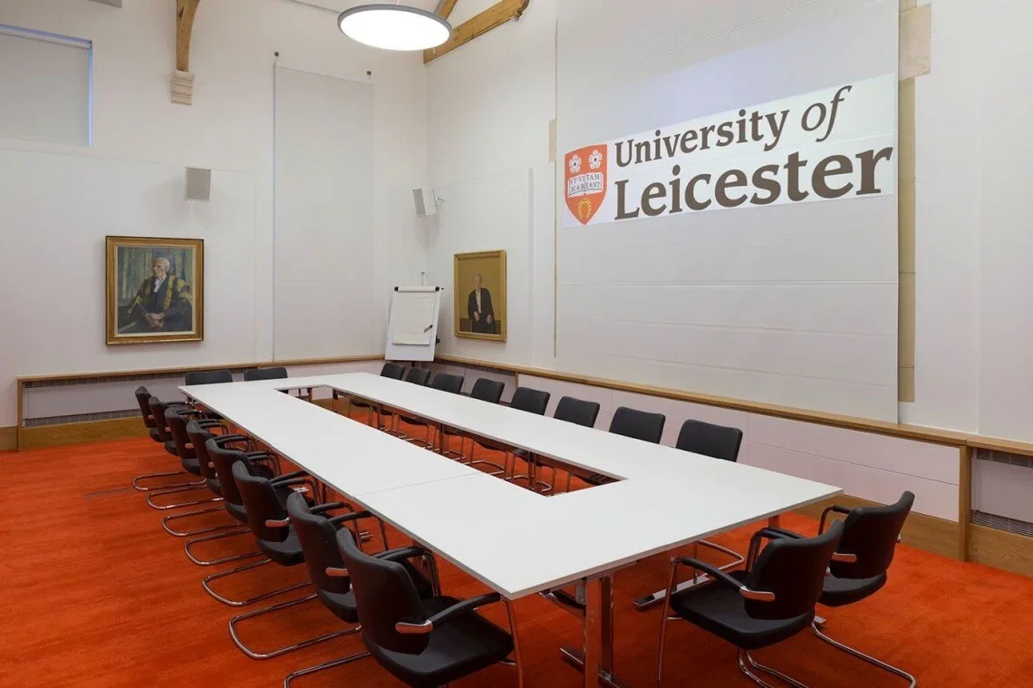 A conference room at the University of Leicester with a U-shaped table, black chairs, and framed portraits on the white walls. A projector screen displays the university logo.
