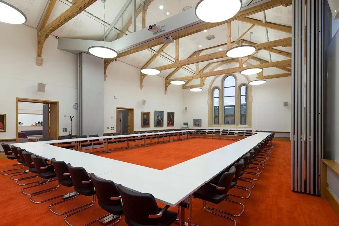 Conference room with U-shaped white table, black chairs, large arched windows, framed artwork on cream-colored walls, and wooden beams on ceiling with circular white lights.