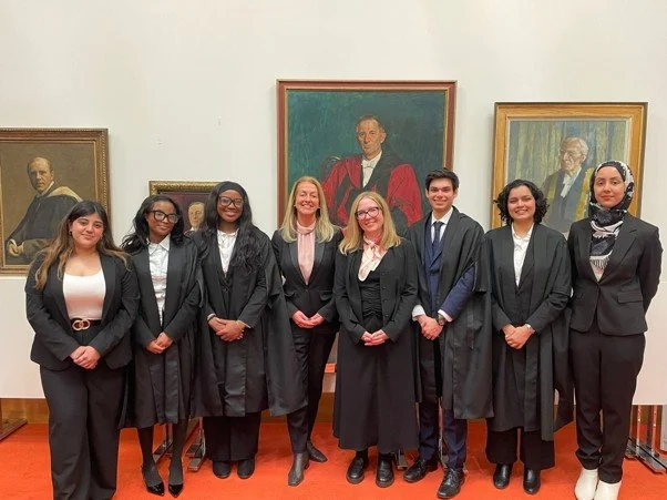 Group of seven diverse people in graduation gowns standing in front of framed portraits in a gallery setting.