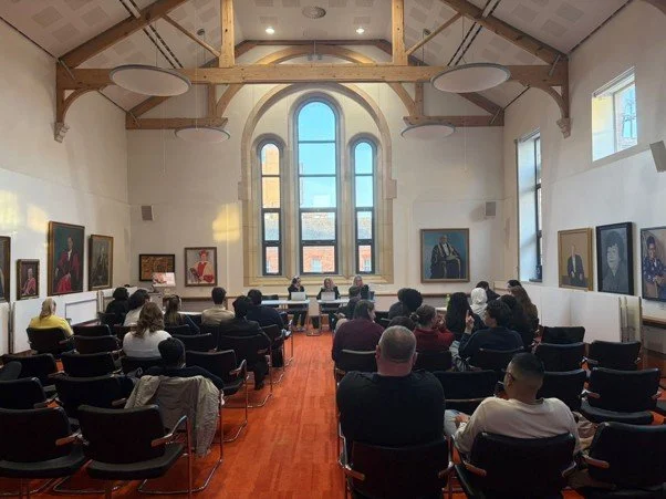 A group of people attending a presentation or lecture in a room with high ceilings, wooden beams, large arched windows, and framed artwork on the walls.