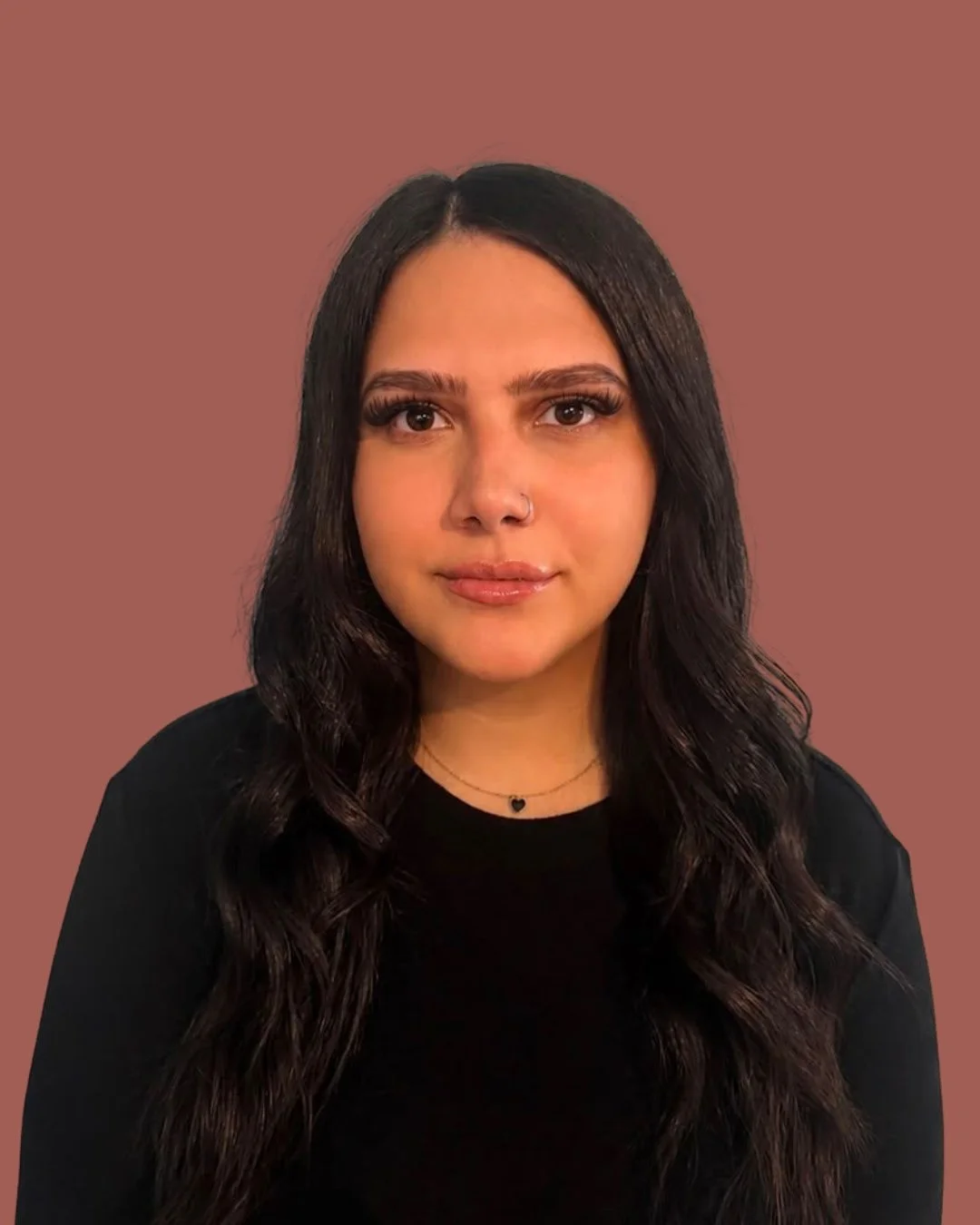 A woman with long dark wavy hair, wearing a black top, a necklace with a heart pendant, and a nose ring, posing against a plain pink background.