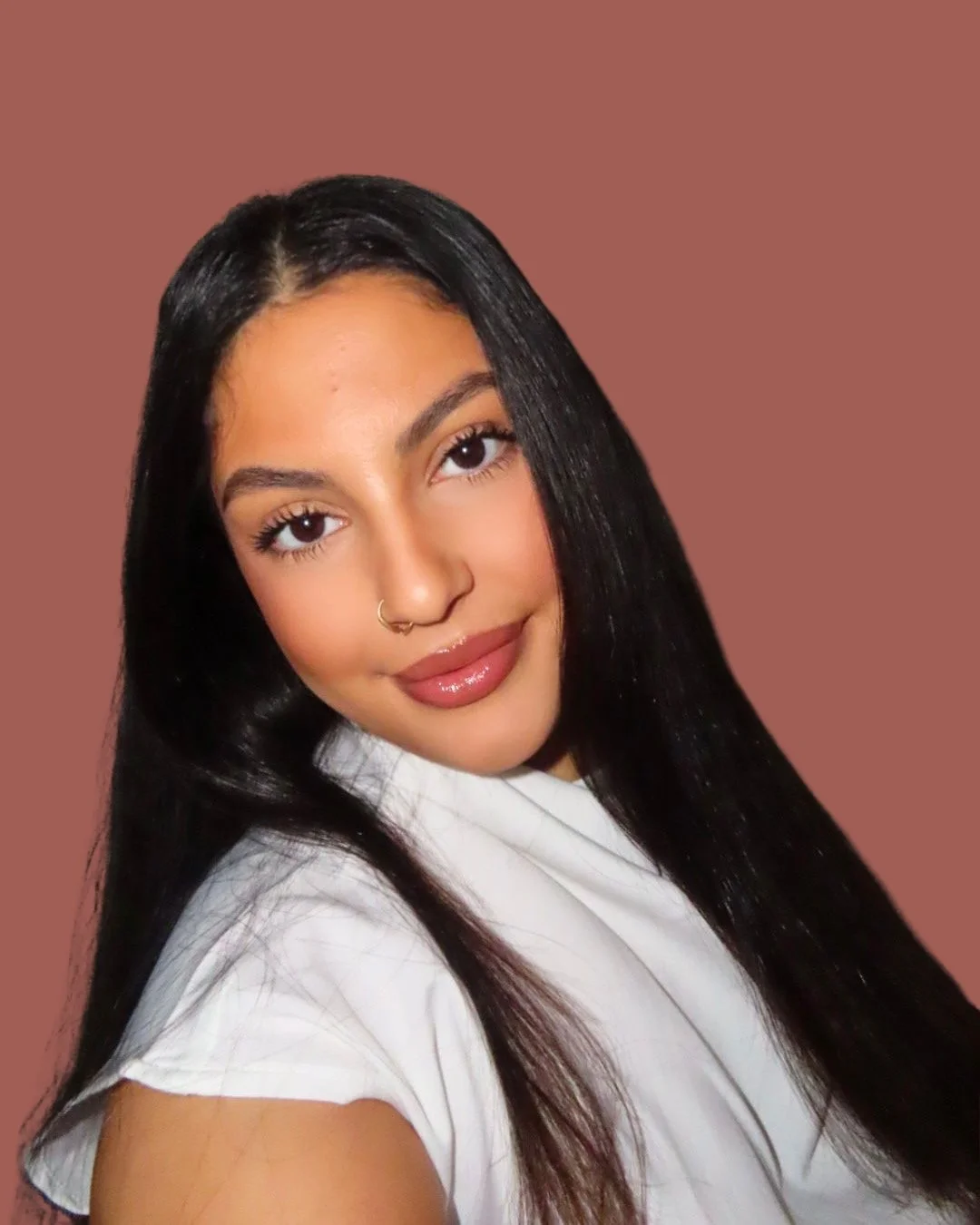Portrait of a woman with long black hair, light brown skin, wearing a white top and a nose ring, smiling with a pinkish-brown background.