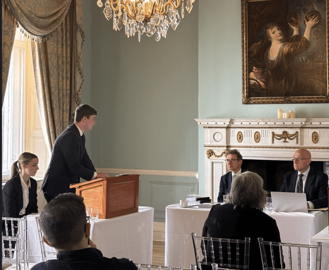 A young man in a dark suit stands at a wooden podium giving a presentation in a formal setting, with three seated people, two men in suits and a woman, listening and looking attentive. The room has ornate gold and white decor, a large chandelier, a fireplace, and a painting of a woman holding two bowls on the wall.