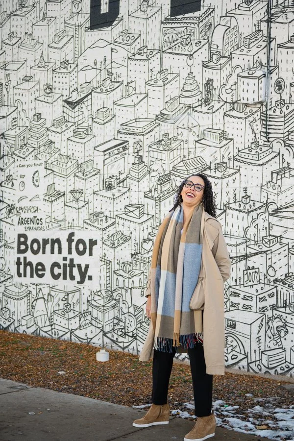 Cherri McKinney laughing outside in a coat in front of a mural that says Born for the city.