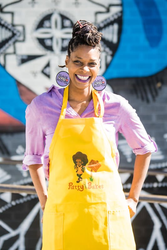 Patty Bakes founder Tiya Trent wearing yellow apron and black lives matter earrings in front of CoCuA restaurant mural