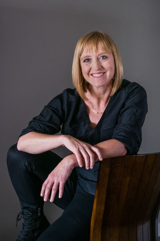 Jill Eelkema, a care manager, wearing a black shirt sits on wood chair for her headshot against a gray wall.