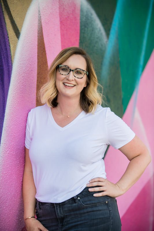 Headshot of Lara Smedley Events wearing white shirt and glasses in front of mural wall in RiNo Denver