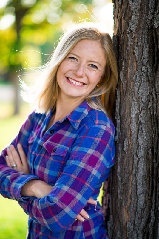 Director Kathryn Jantz posing for a personal portrait against a tree wearing a plaid blue shirt with light in her hair