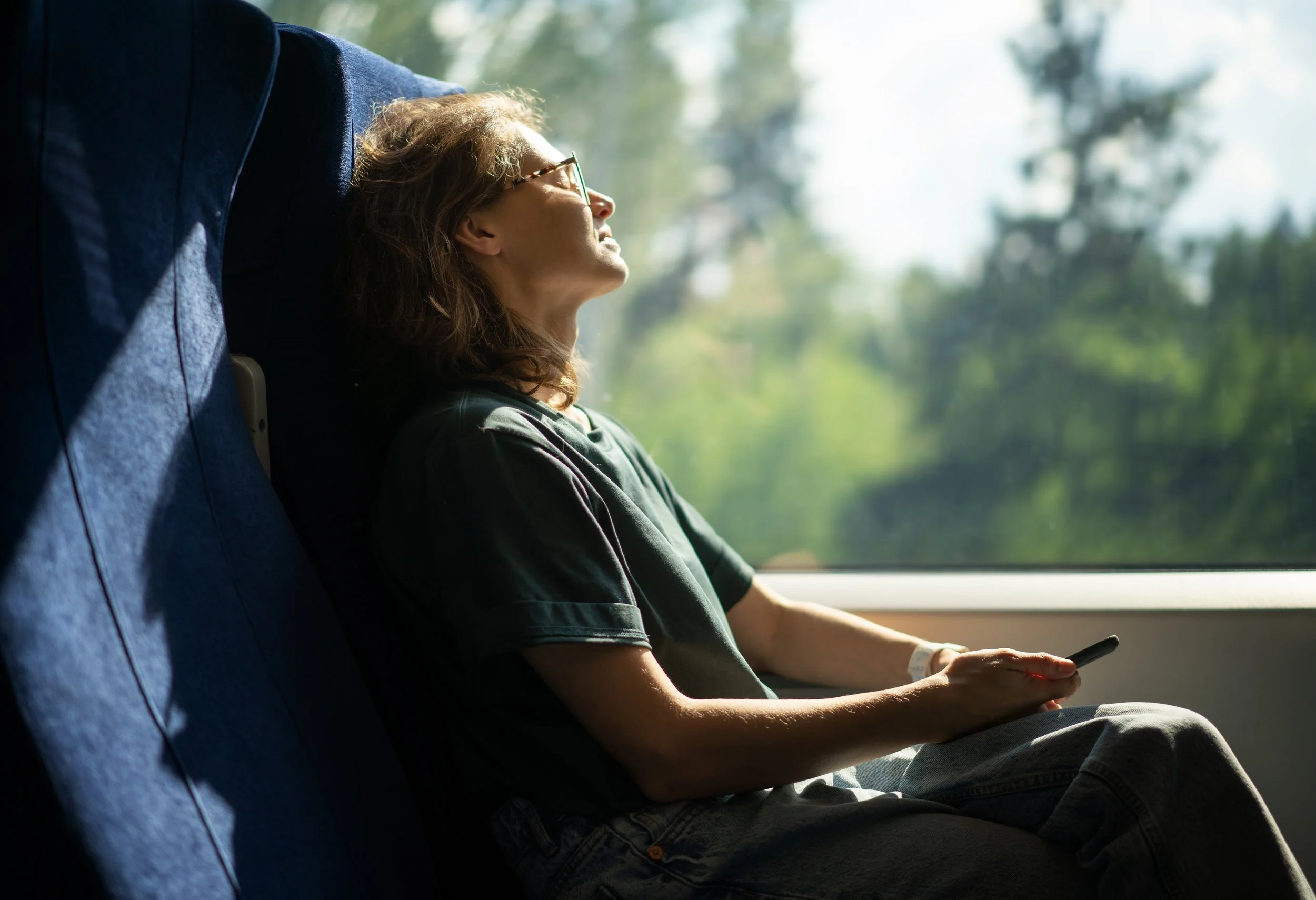 Woman sitting on a train, head laid back on the seat, eyes closed, holding her phone and enjoying the sun coming through the window.