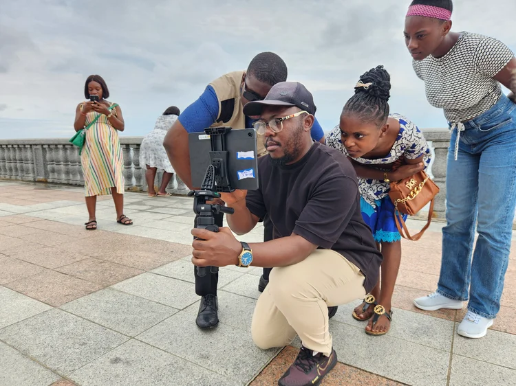 A trainer films with a tablet-based camera rig on a tripod while community members look on.