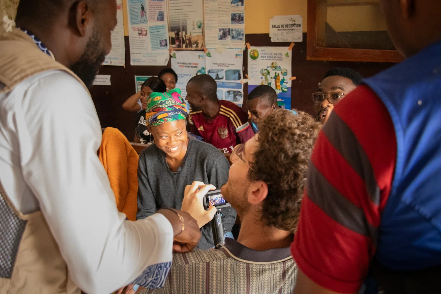 A videographer films a community member mid-interview during a screening session, surrounded by health education posters.
