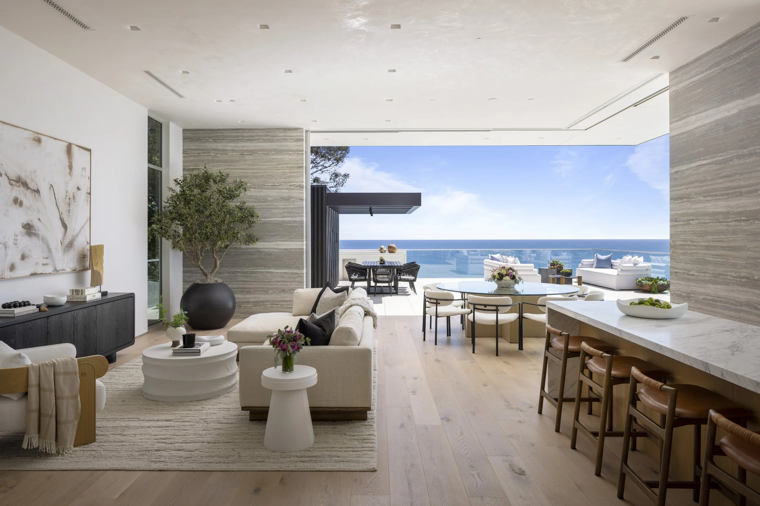Modern living room with large sliding glass doors leading to balcony with ocean view, beige sofa with black pillows, marble countertops, wooden chairs, potted tree, and artwork on wall.
