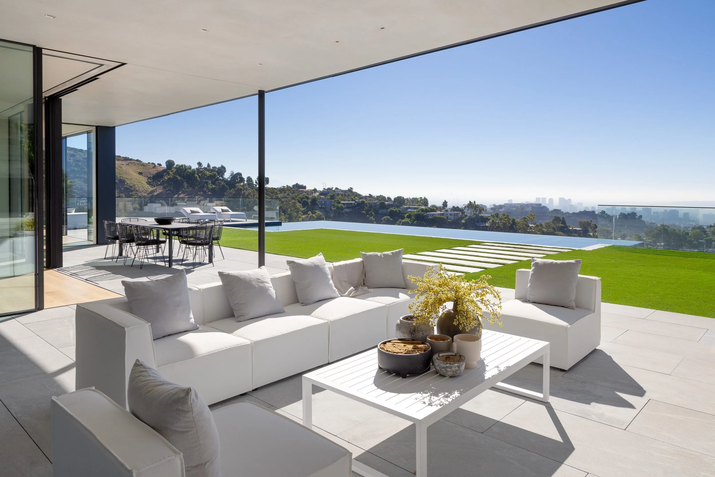 Modern outdoor patio with white sectional sofa and a coffee table with vases and flowers, overlooking a green lawn and a city skyline in the distance under a clear blue sky.