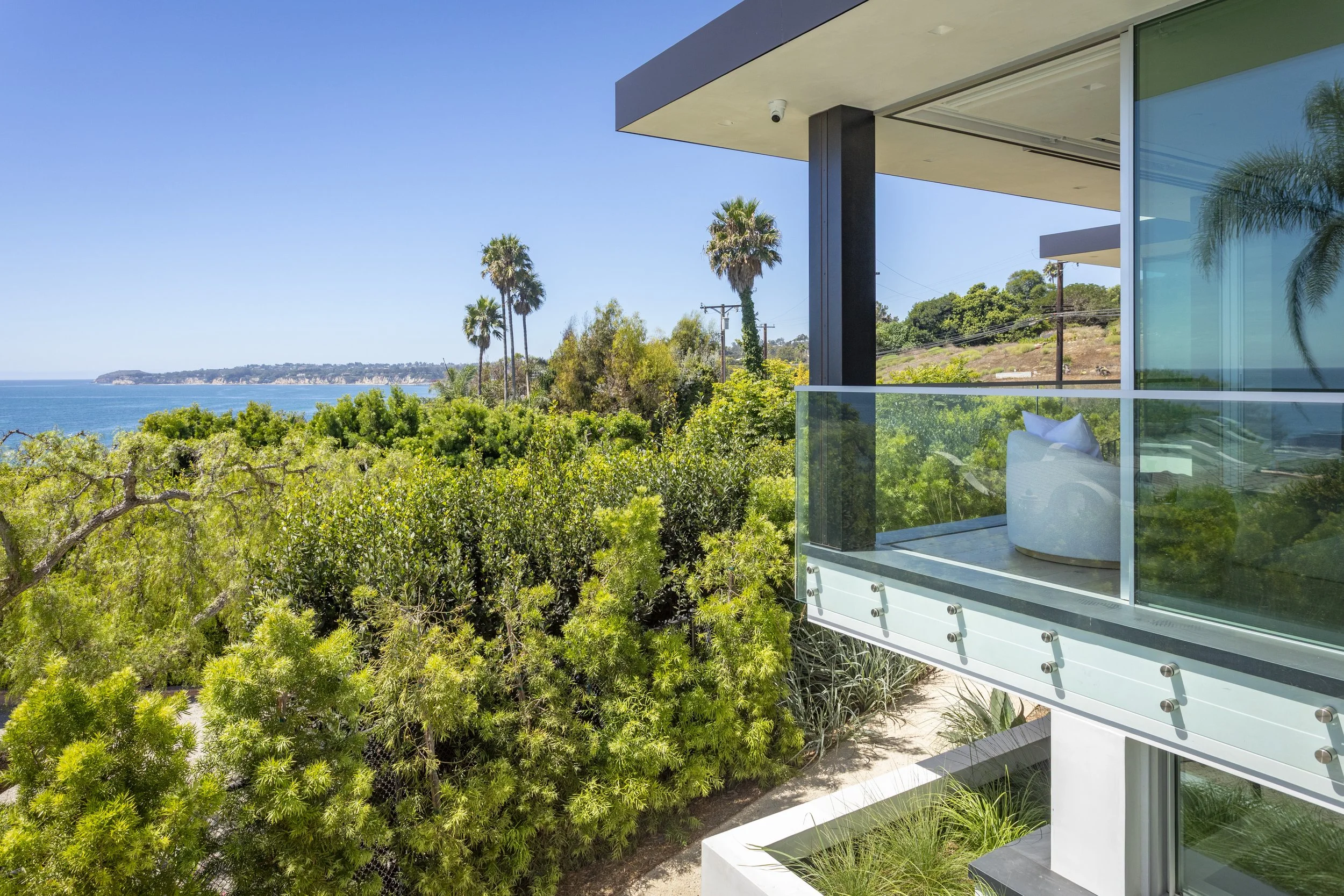 A modern house with a glass balcony overlooking lush green trees and the ocean under a clear blue sky.