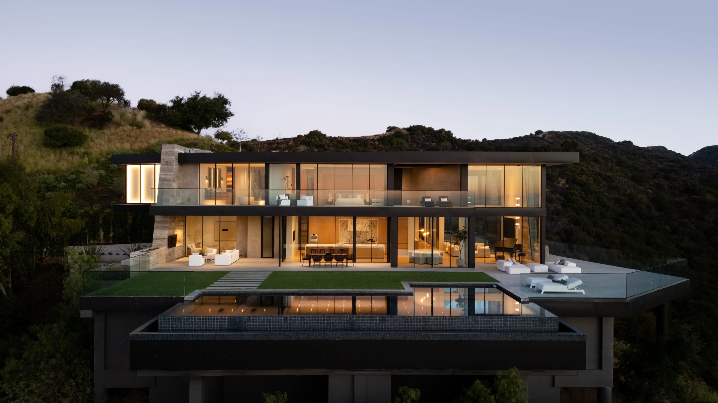 A modern multi-story house with glass walls and an infinity pool, located on a hillside at dusk.