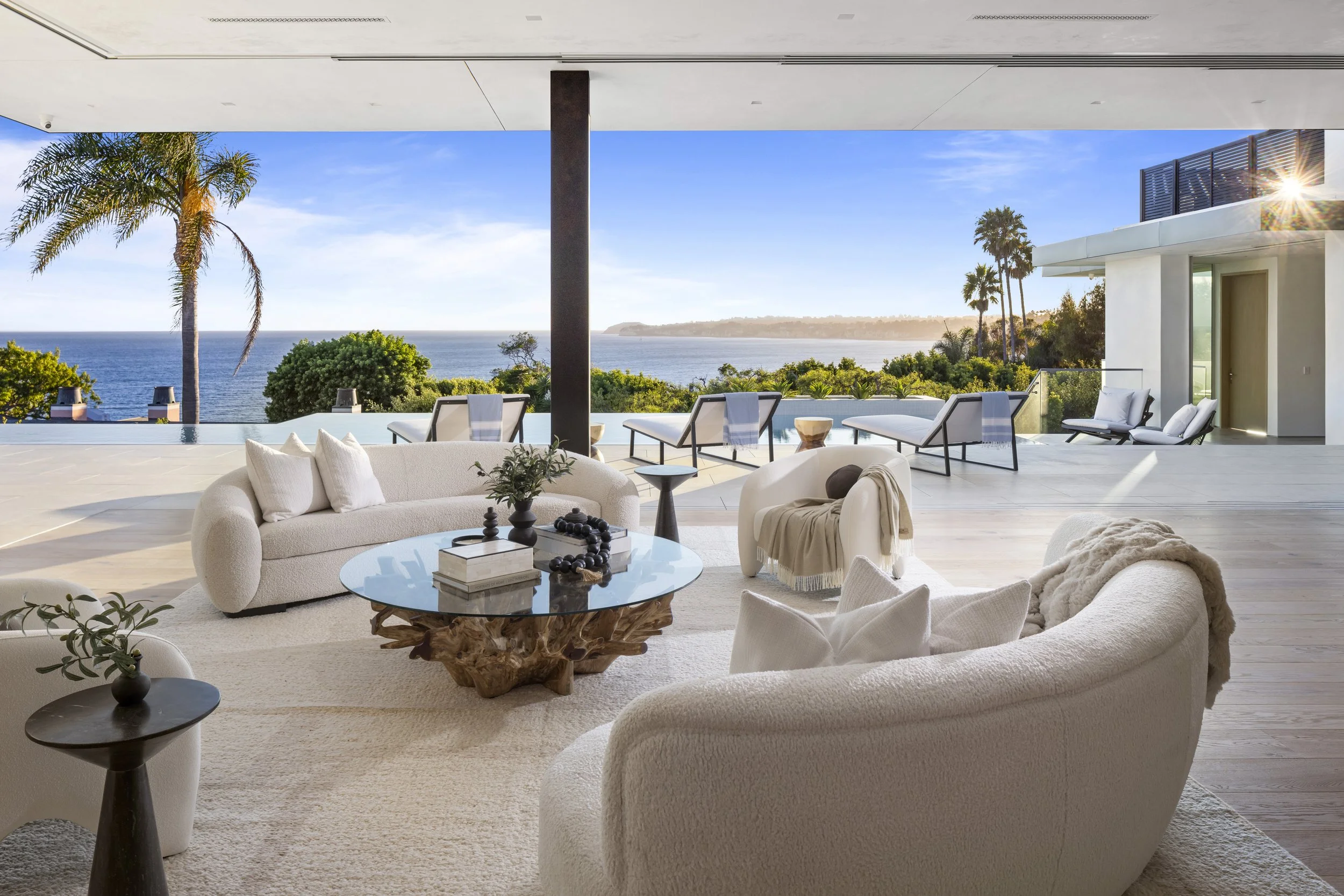 Open living room with large windows showing a view of the ocean, palm trees, and a bright blue sky, decorated with white sofas, a glass-topped coffee table, and outdoor lounge chairs.