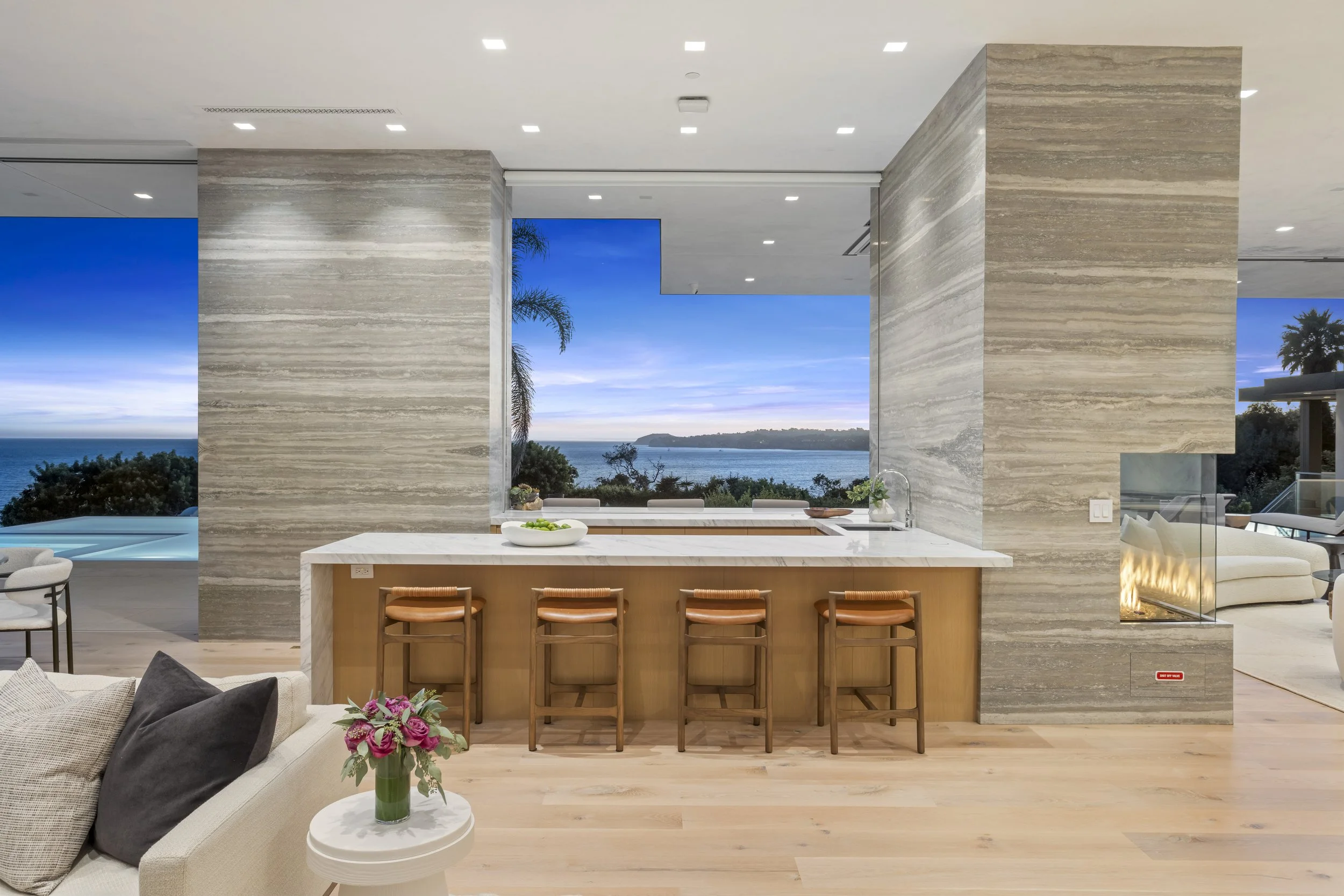 Modern open-concept living space with a view of the ocean through large windows, featuring a kitchen with a marble island, four bar stools, a patio with a pool, and interior seating areas with a fireplace and flowers.