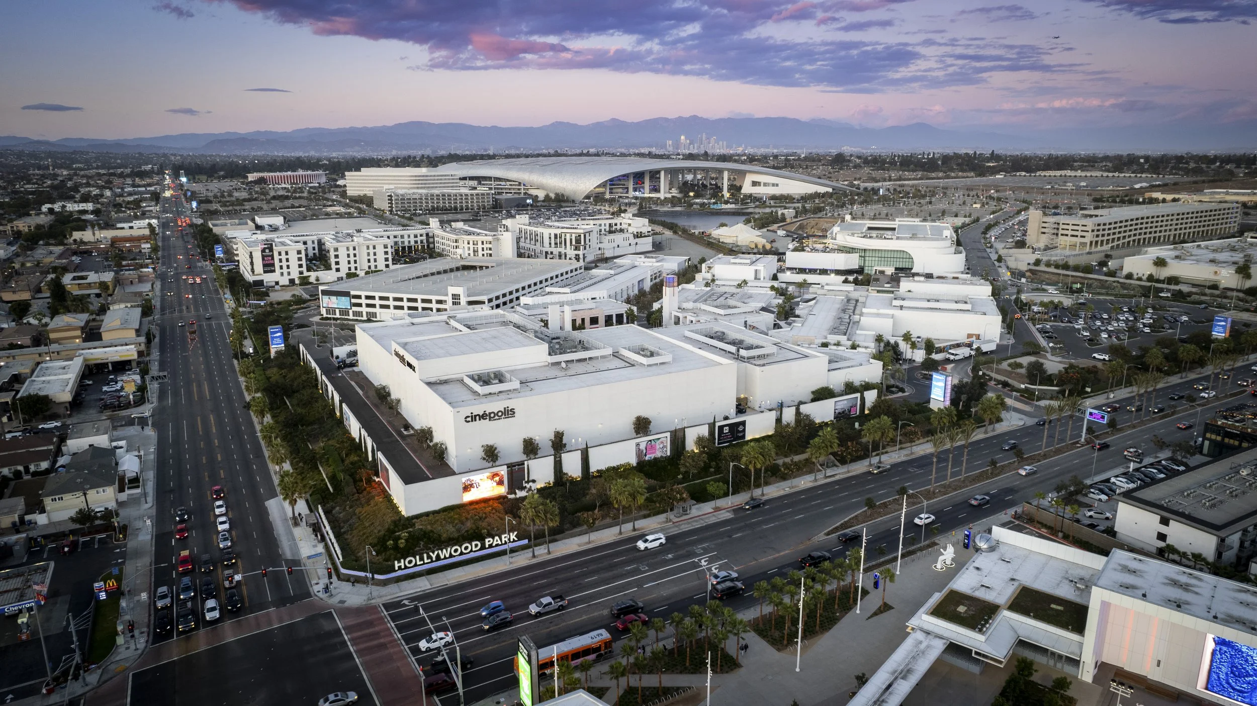 Aerial view of Hollywood Park in Los Angeles, California with buildings, streets, parking lots, and the SoFi Stadium in the background during sunset.