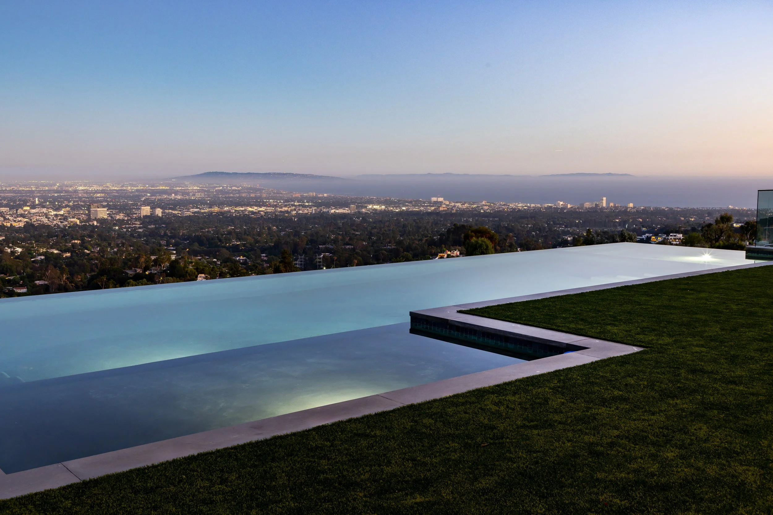 Luxury infinity pool with city and ocean view at dusk, featuring a glass wall and grassy area.