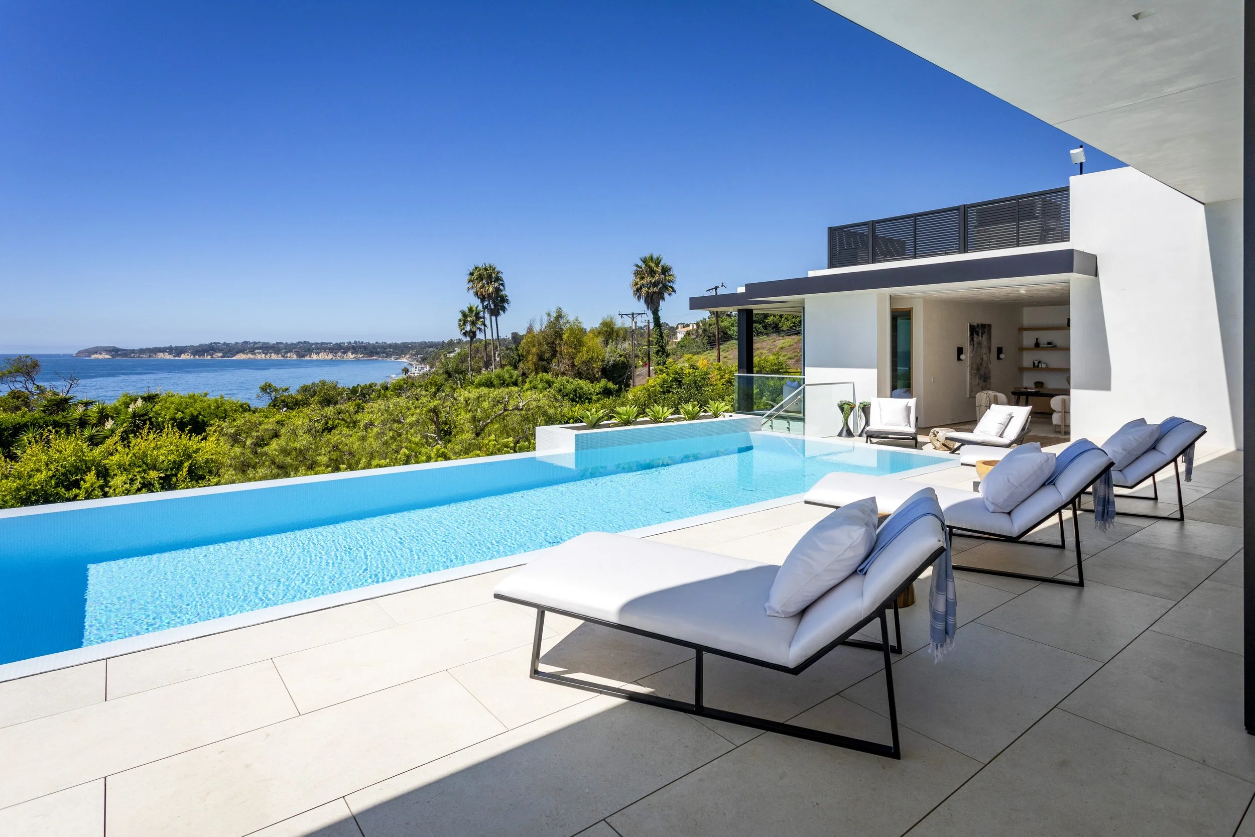 Modern house with an infinity pool overlooking a scenic ocean view, with white lounge chairs on the patio.