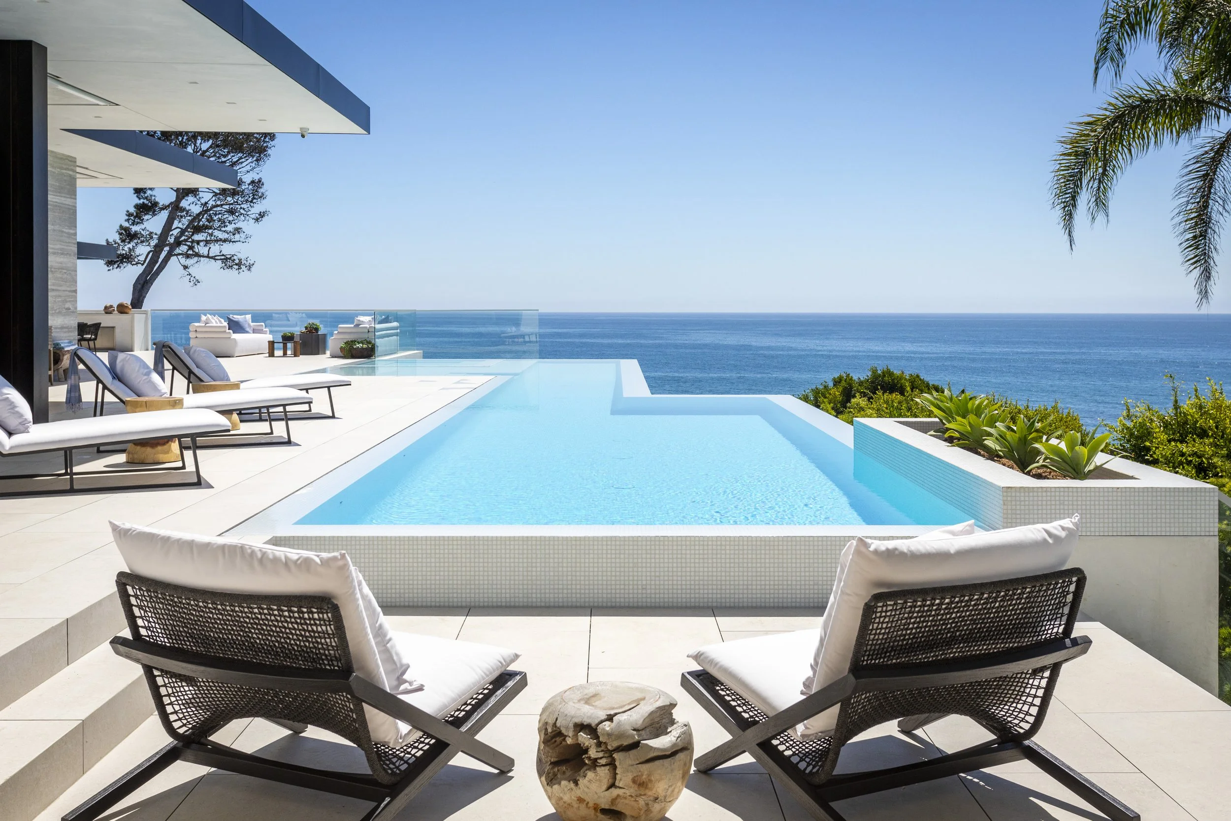 Luxury infinity pool overlooking the ocean on a sunny day, with outdoor lounge chairs and green plants.