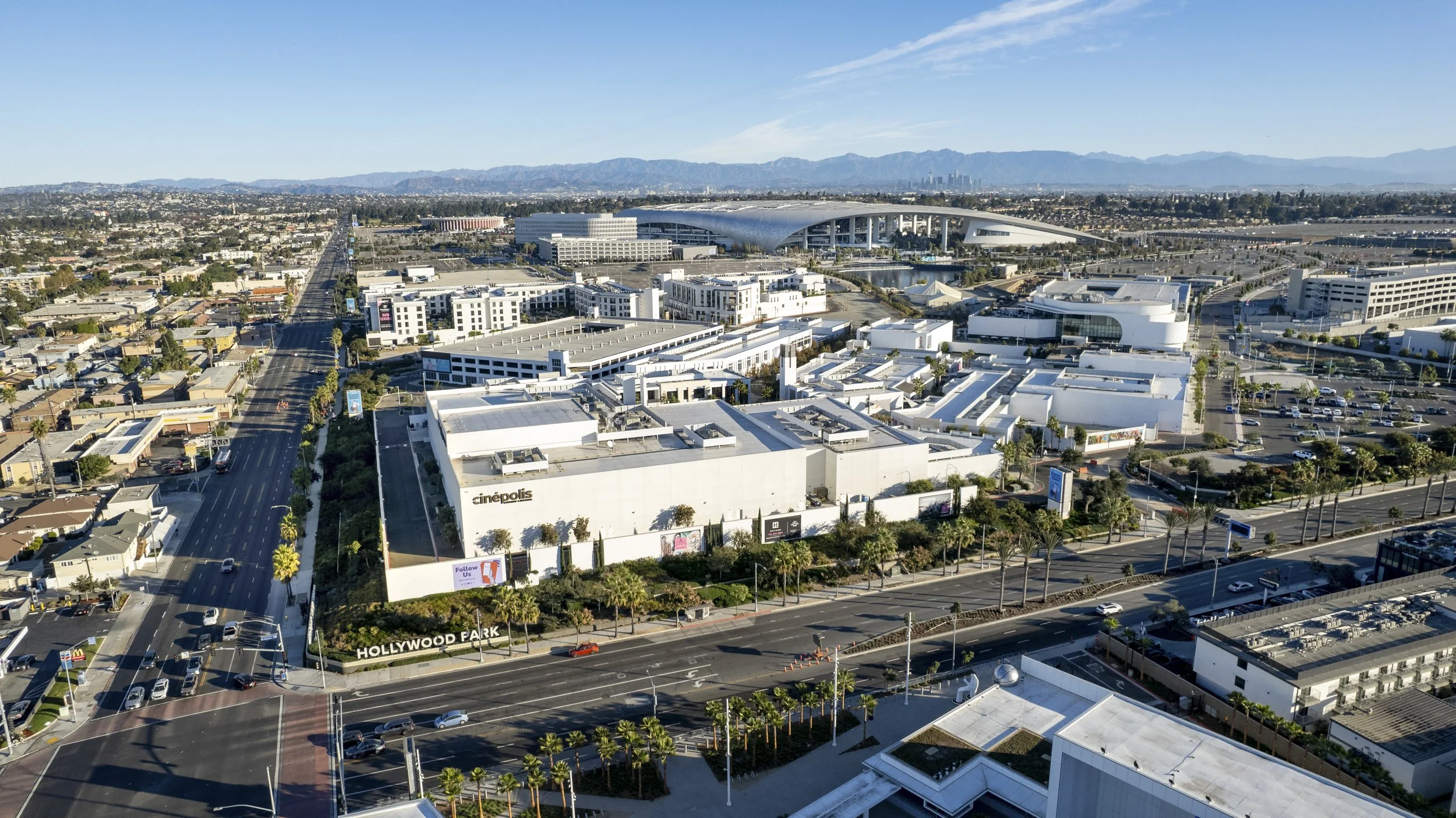 Aerial view of Hollywood Park area in Los Angeles, California, showing white commercial buildings, parking lots, streets lined with palm trees, and mountains in the background under a clear blue sky.