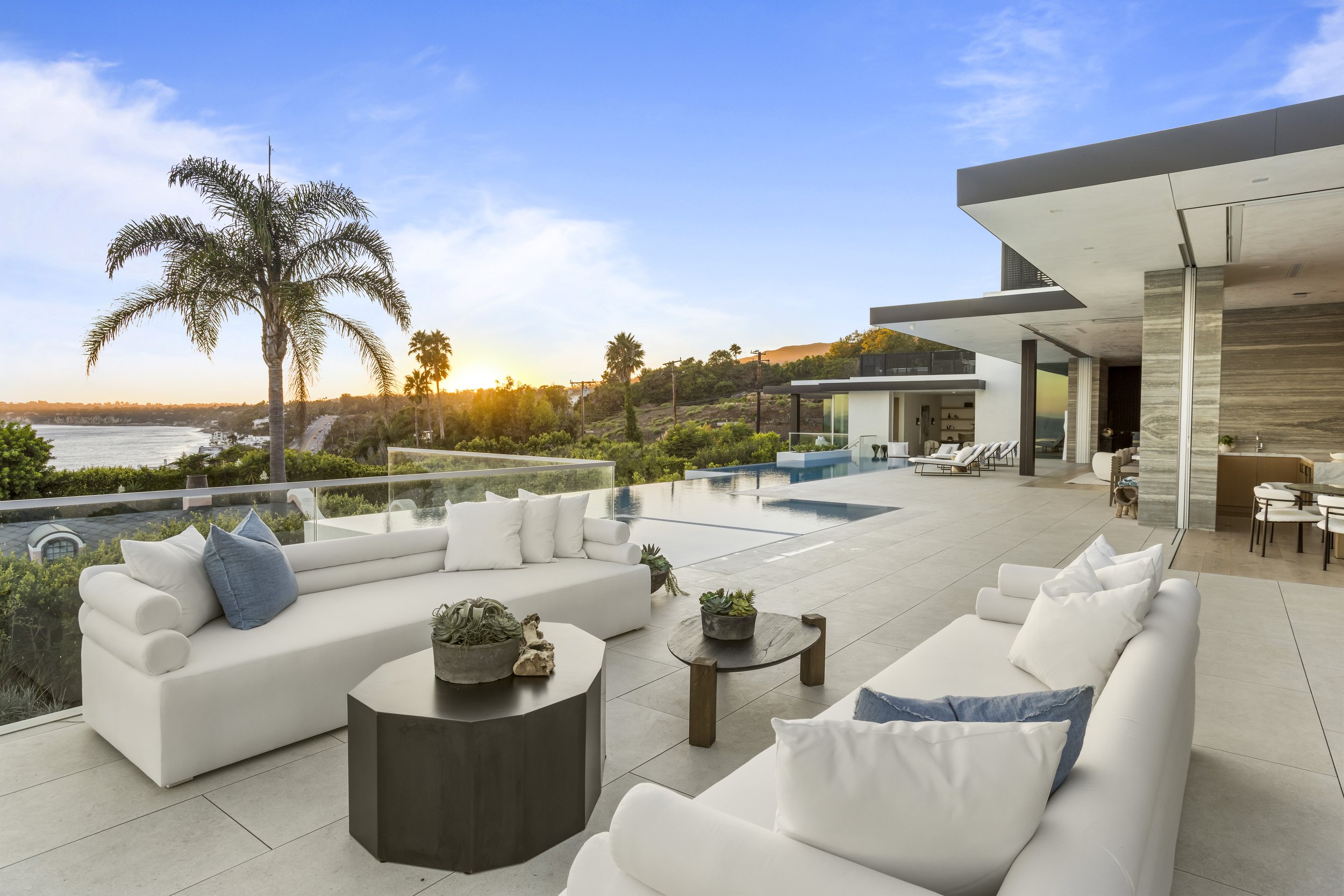 Modern outdoor patio with white sofas, small tables, and potted plants, overlooking a pool and a scenic view of a river, palm trees, and a sunset.