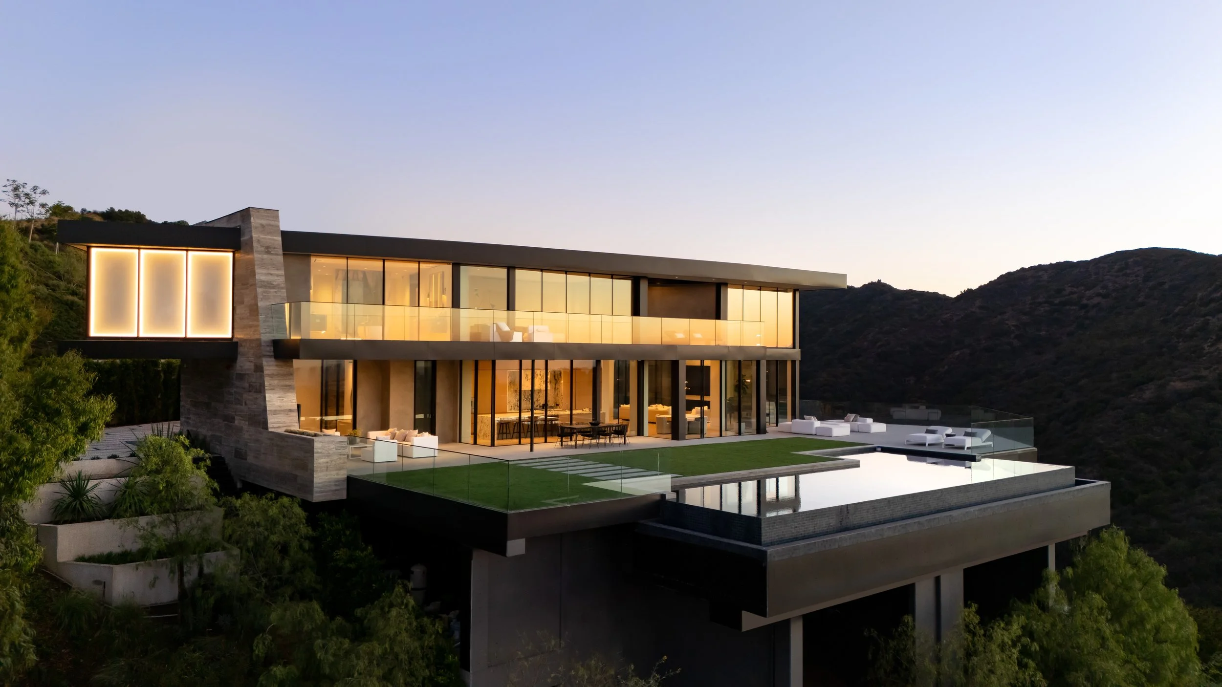 Modern house on hillside with large glass windows, outdoor patio, and infinity pool, surrounded by green trees and mountain landscape at sunset.