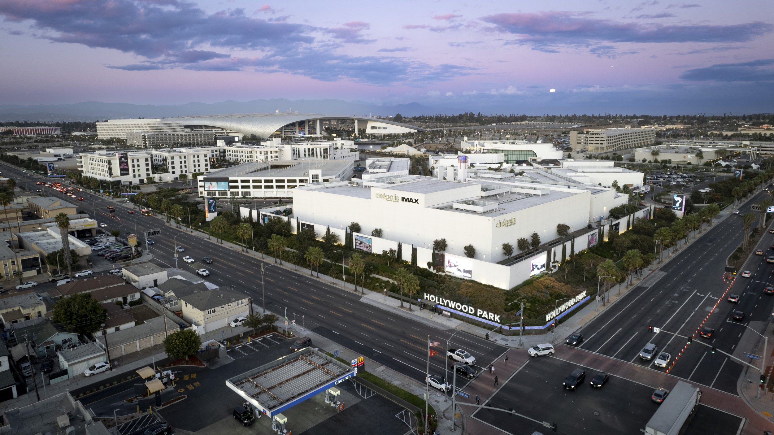An aerial view of Hollywood Park, including the Sofi Stadium in the background, nearby shopping and parking areas, and surrounding streets during dusk with a partly cloudy sky.