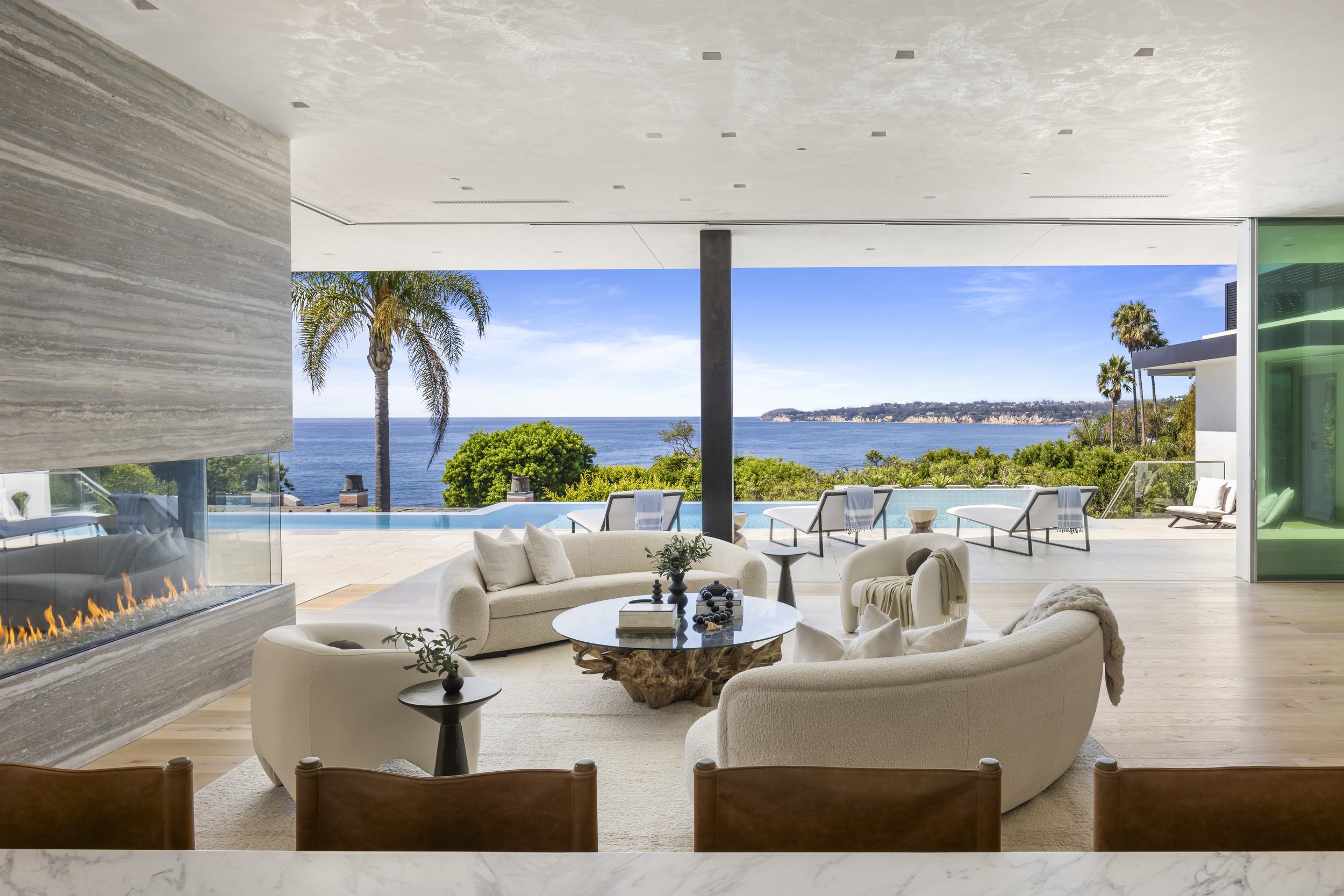 Modern living room with white sofa and armchairs, glass coffee table, and fireplace, overlooking an outdoor pool and ocean view with palm trees.