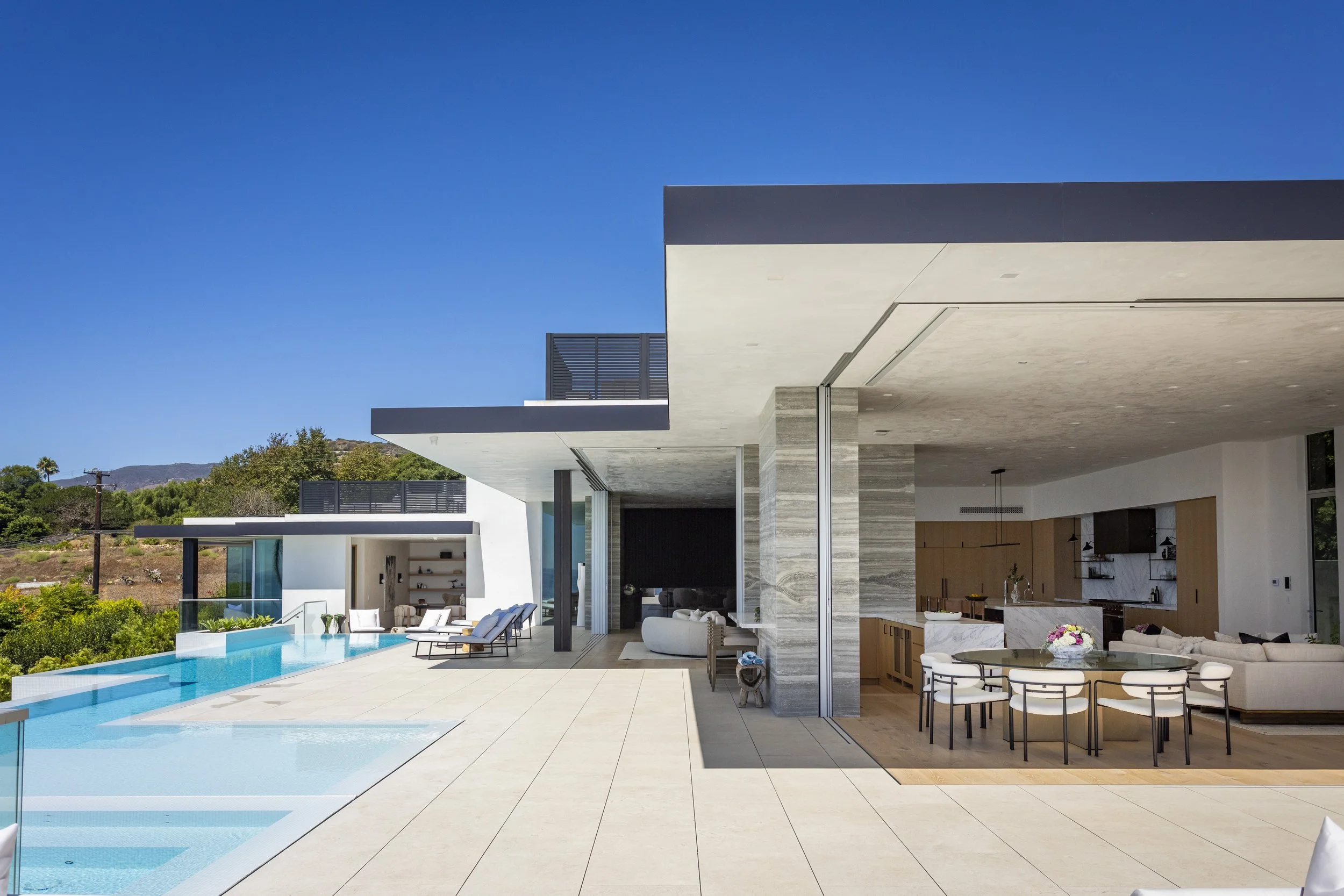 Modern house with an outdoor pool, lounge chairs, and an open interior with dining area and living room, under a clear blue sky.