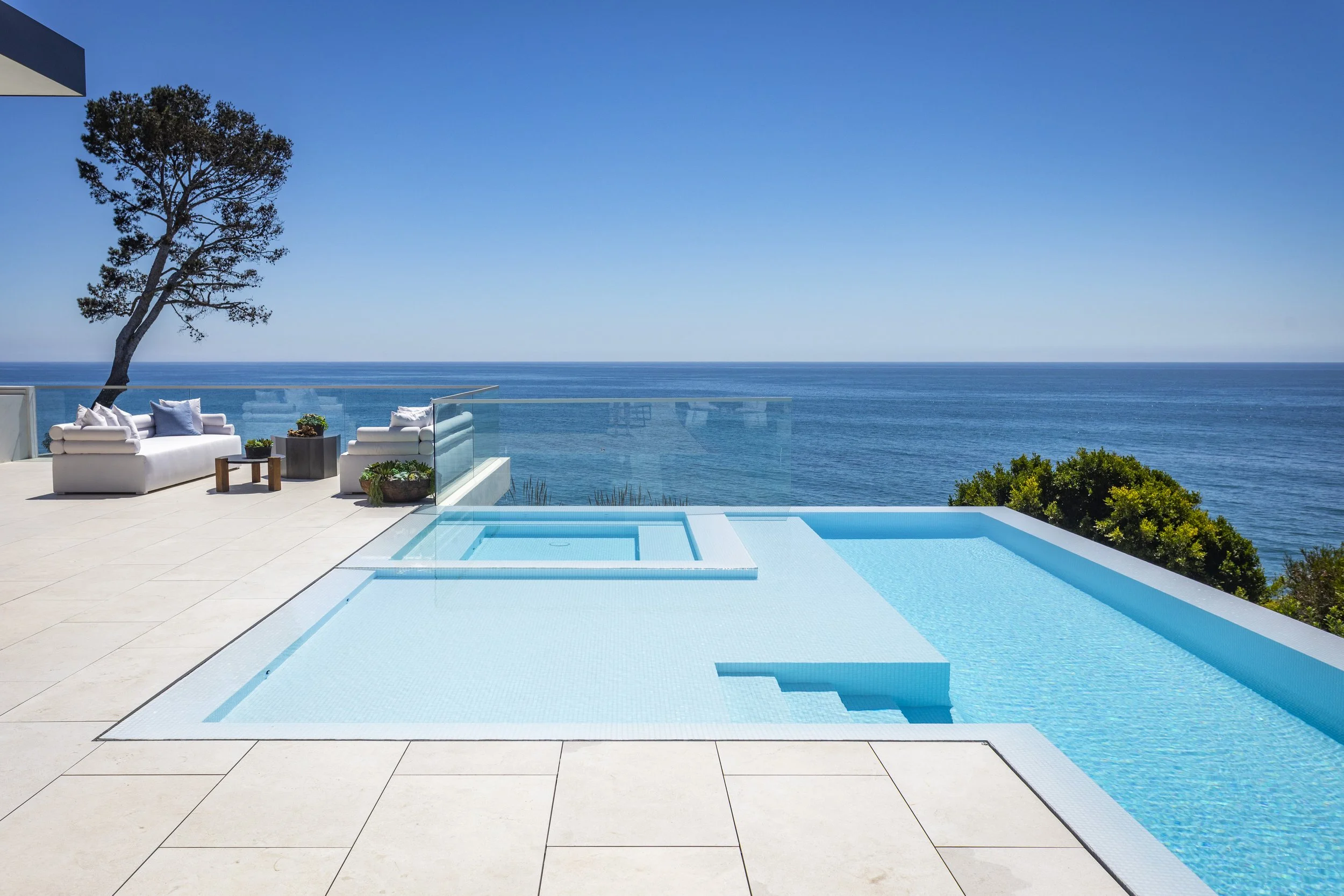 Luxury oceanfront pool with clear blue water, outdoor patio with white seating, and a view of the ocean and sky.