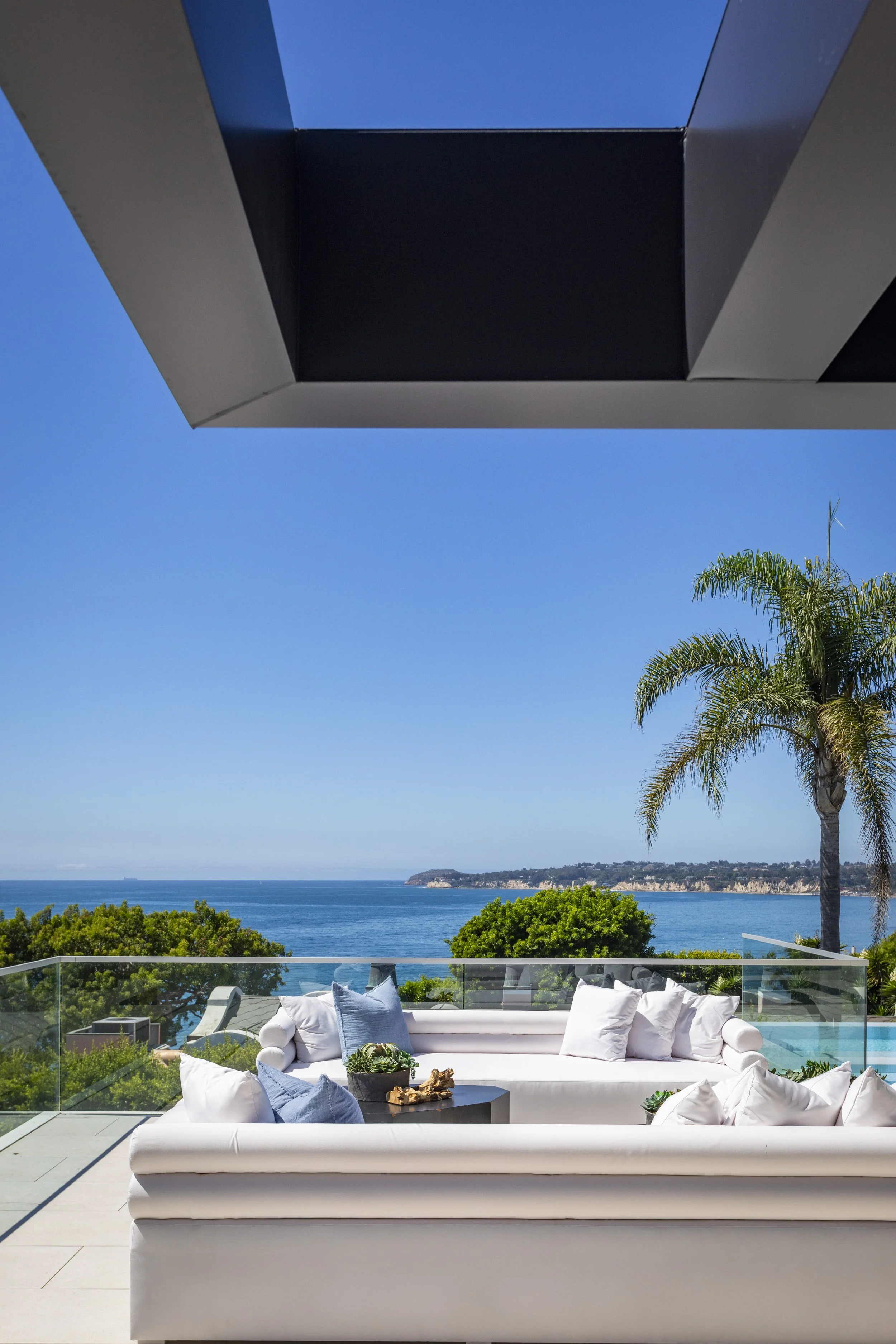 Ocean view from a balcony with white outdoor sofas and blue pillows, glass railing, palm tree, clear blue sky.