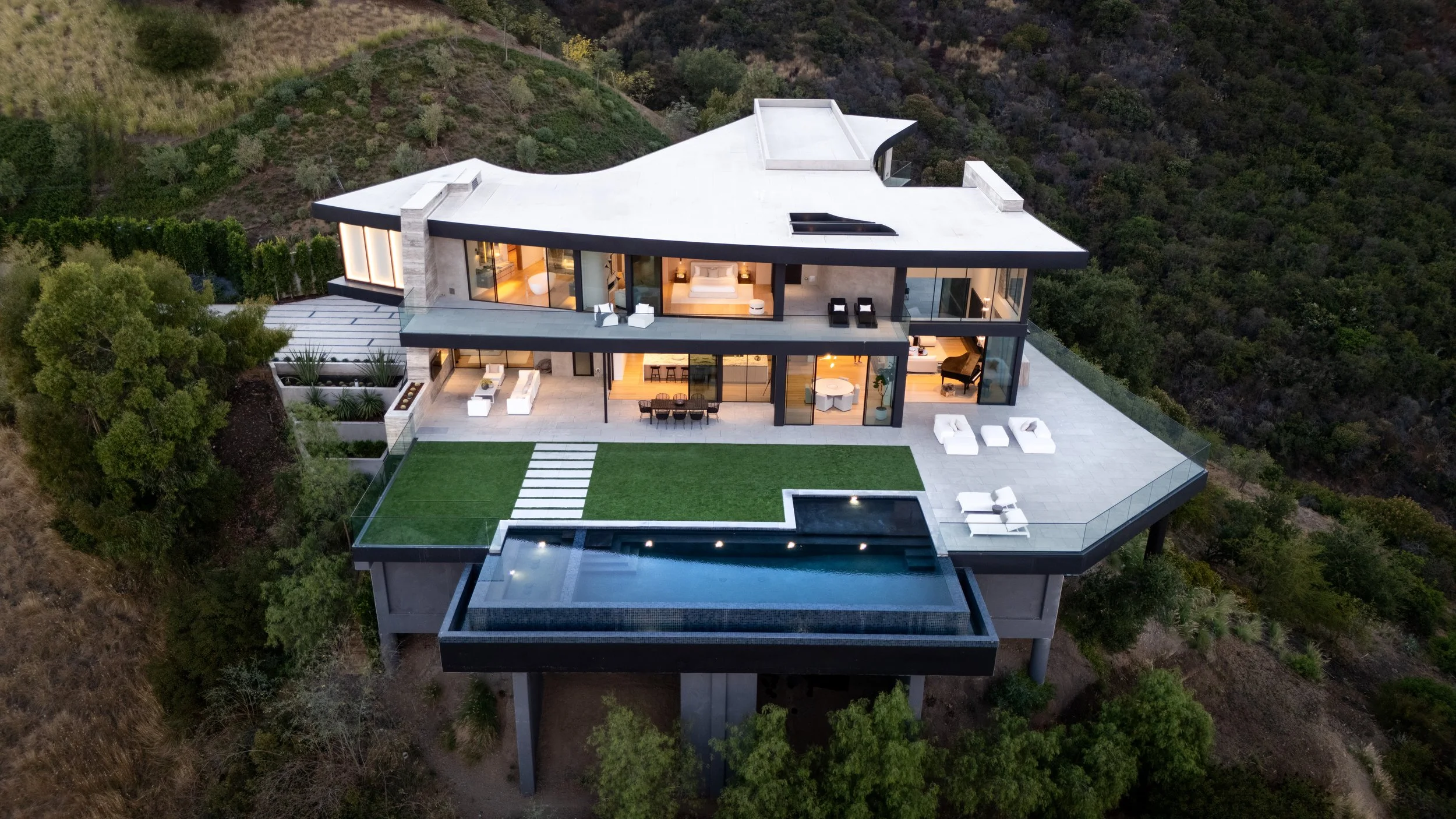 A modern multi-level house built on a hillside with extensive glass walls, a large balcony, outdoor seating, and a sleek pool underneath, surrounded by trees and greenery.