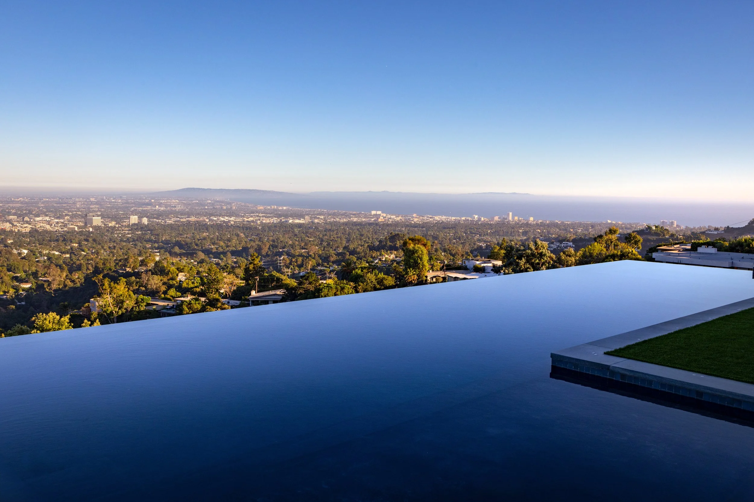 A backyard with a modern infinity pool overlooking a cityscape and distant hills, with clear blue sky above.
