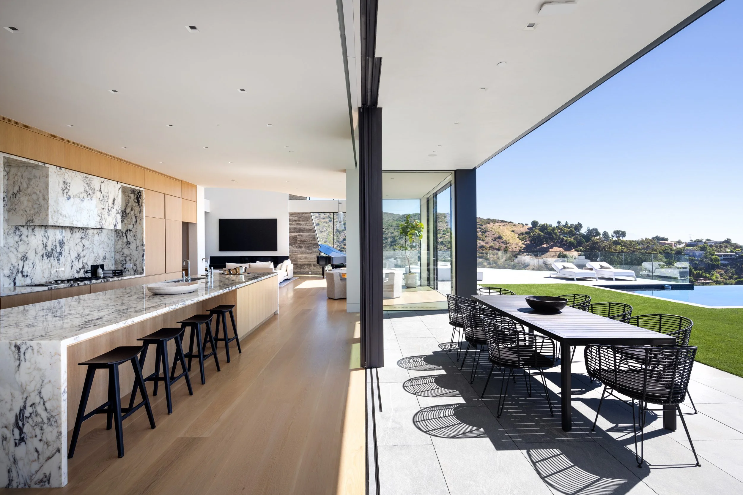 Modern open-concept living space with a marble kitchen island, bar stools, and a view of outdoor patio and landscape with a pool.