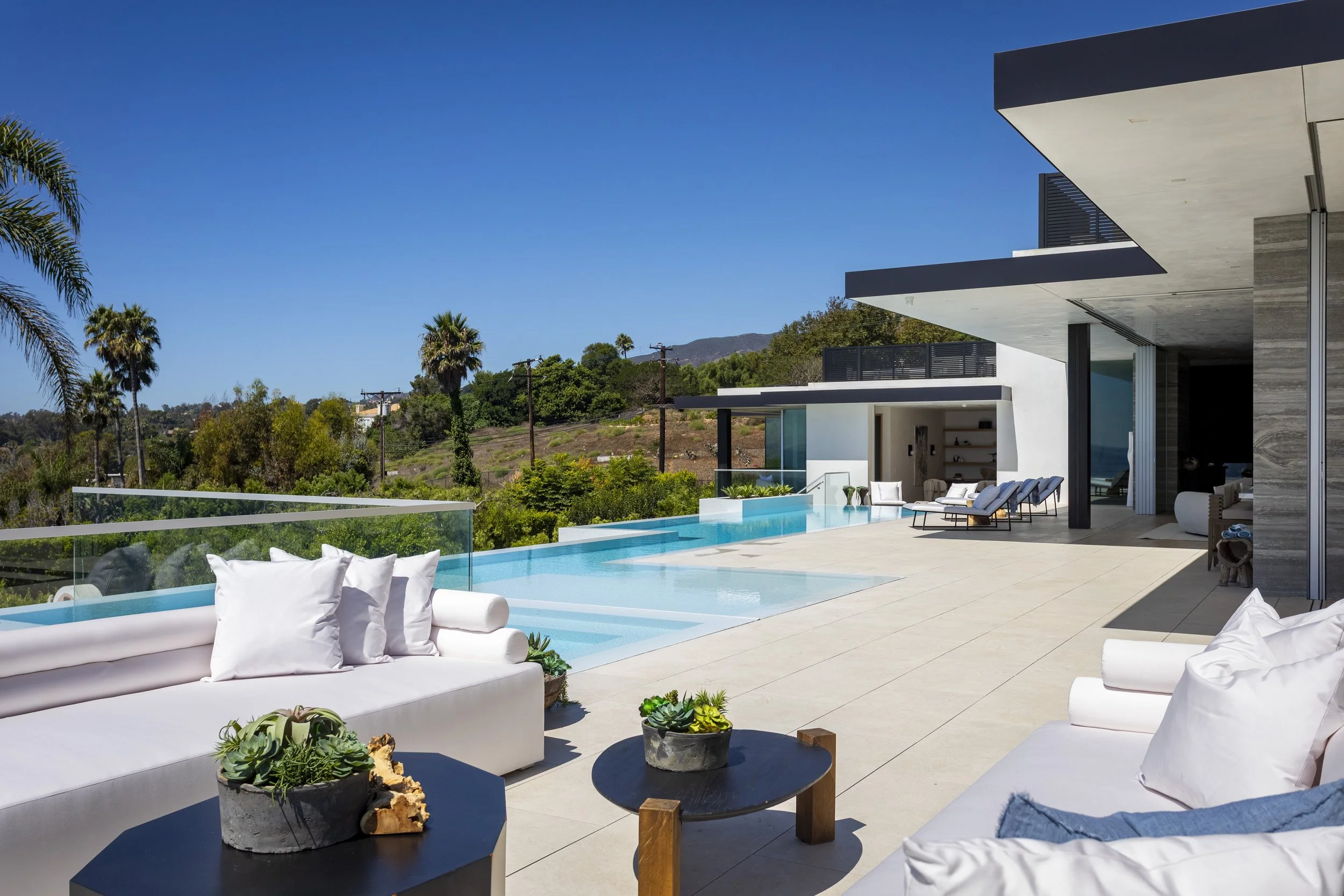Modern house with a pool and outdoor seating area, with a view of palm trees and hills under a blue sky.