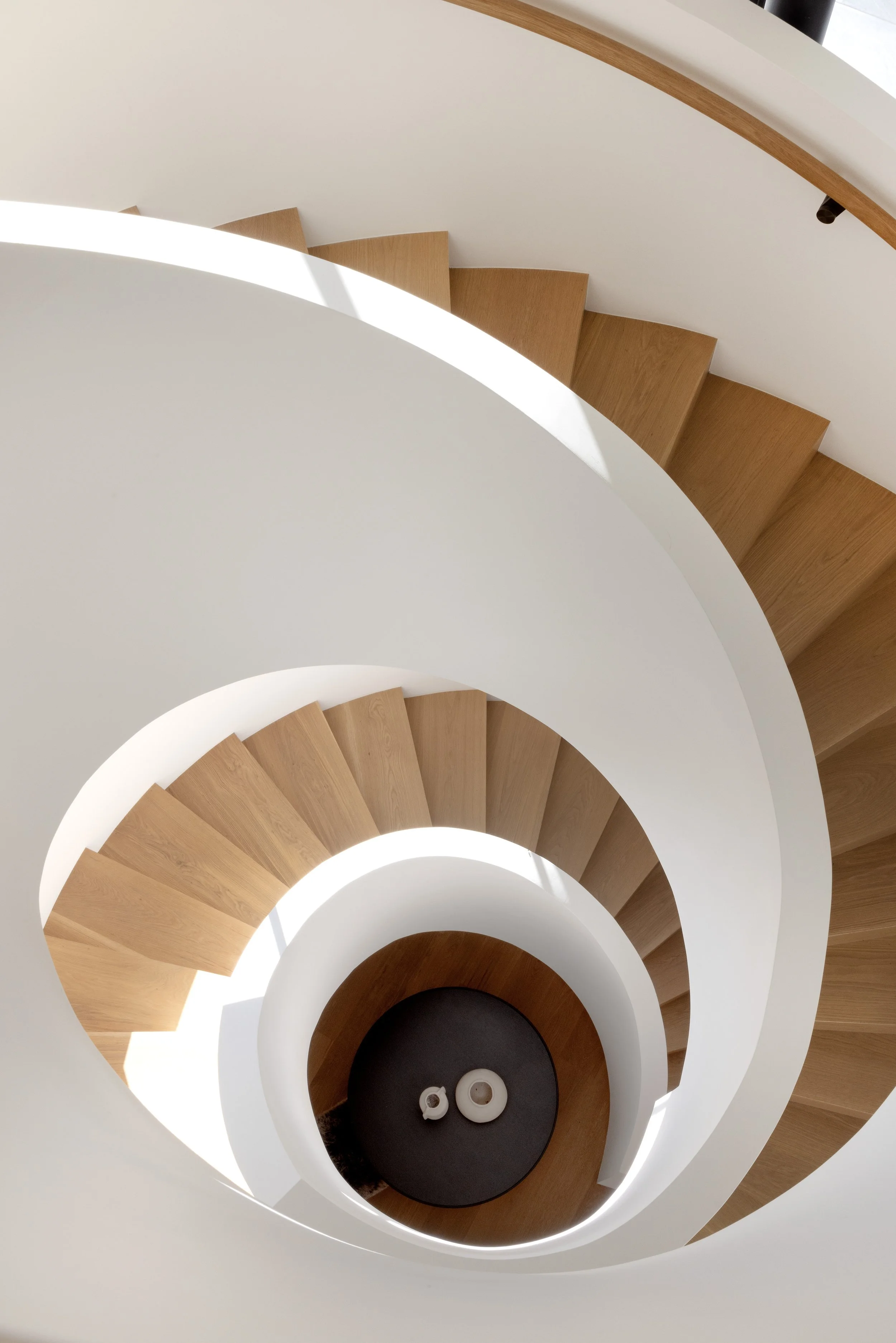 Top-down view of a modern spiral staircase with wooden steps and white sides, winding down to a circular black floor with decorative objects.