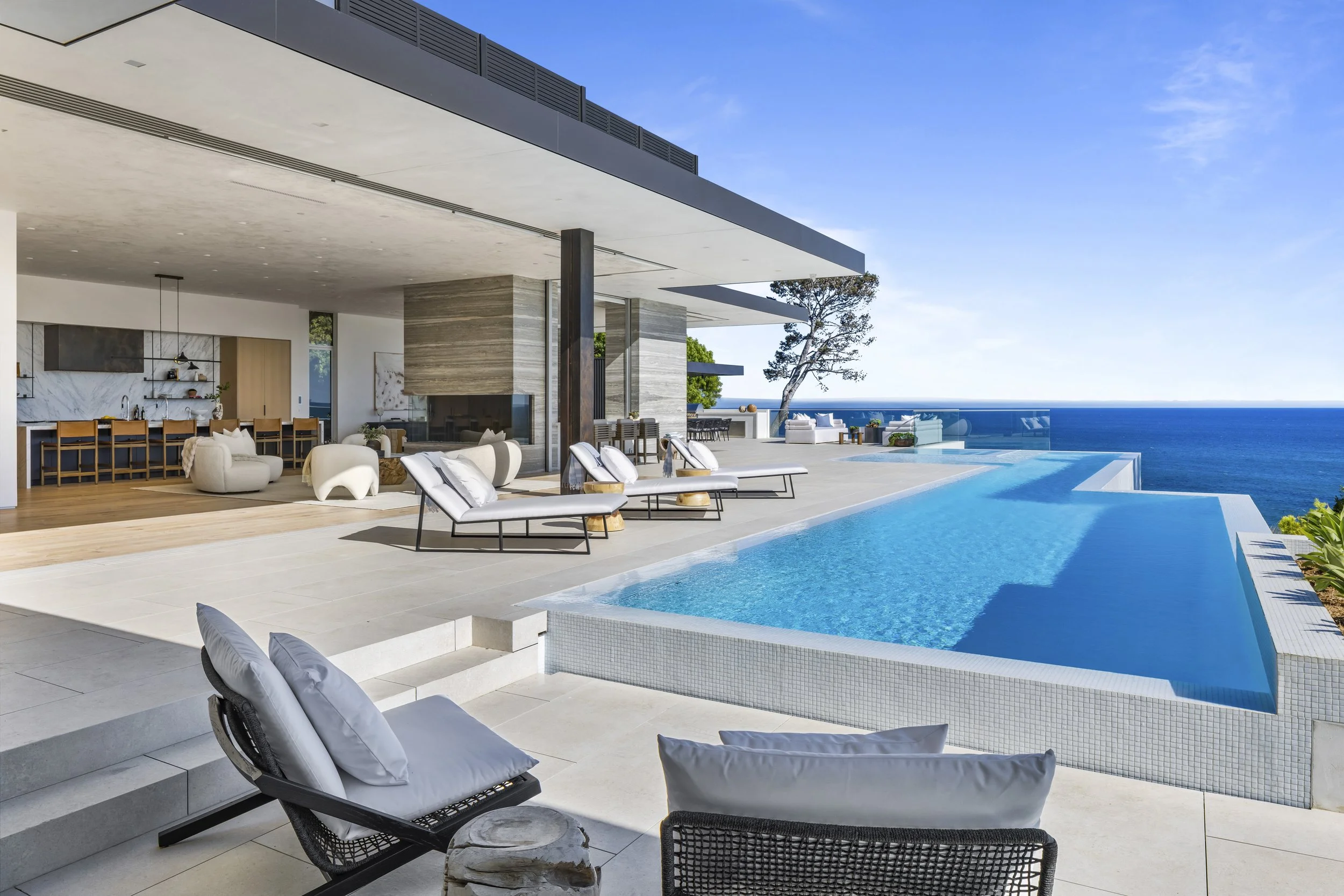 Modern house with outdoor pool overlooking the ocean, featuring lounge chairs, seating area, and a patio with views of the sea and sky.
